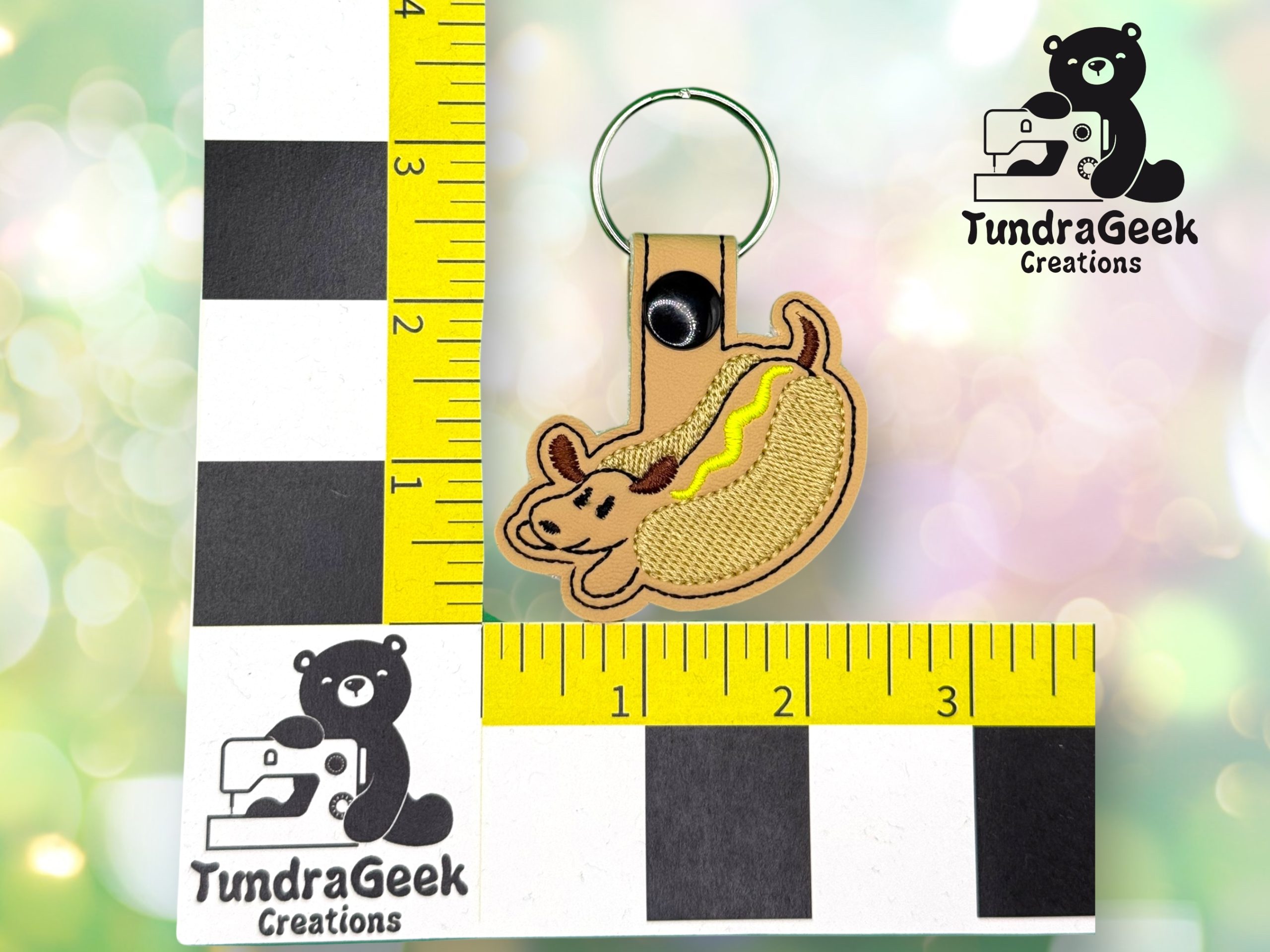 A keychain shaped like a hot dog with a tan bun and yellow mustard, displayed on a ruler grid showing its size, with the TundraGeek Creations logo in the background.