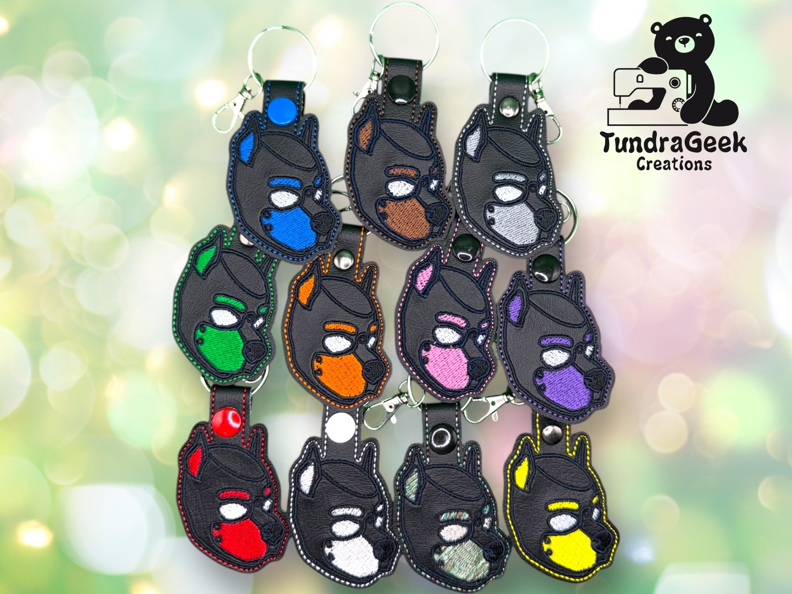 Twelve black dog head keychains with colorful eye and ear accents are arranged neatly; each has a different color and features a metal key ring or clasp. TundraGeek Creations logo is in the corner.