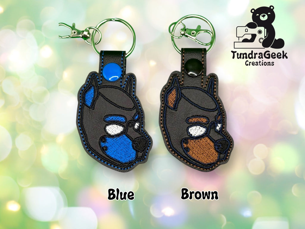Two embroidered keychains shaped like dog heads, one with blue accents and one with brown accents, displayed on a blurred, colorful background. TundraGeek Creations logo is in the corner.