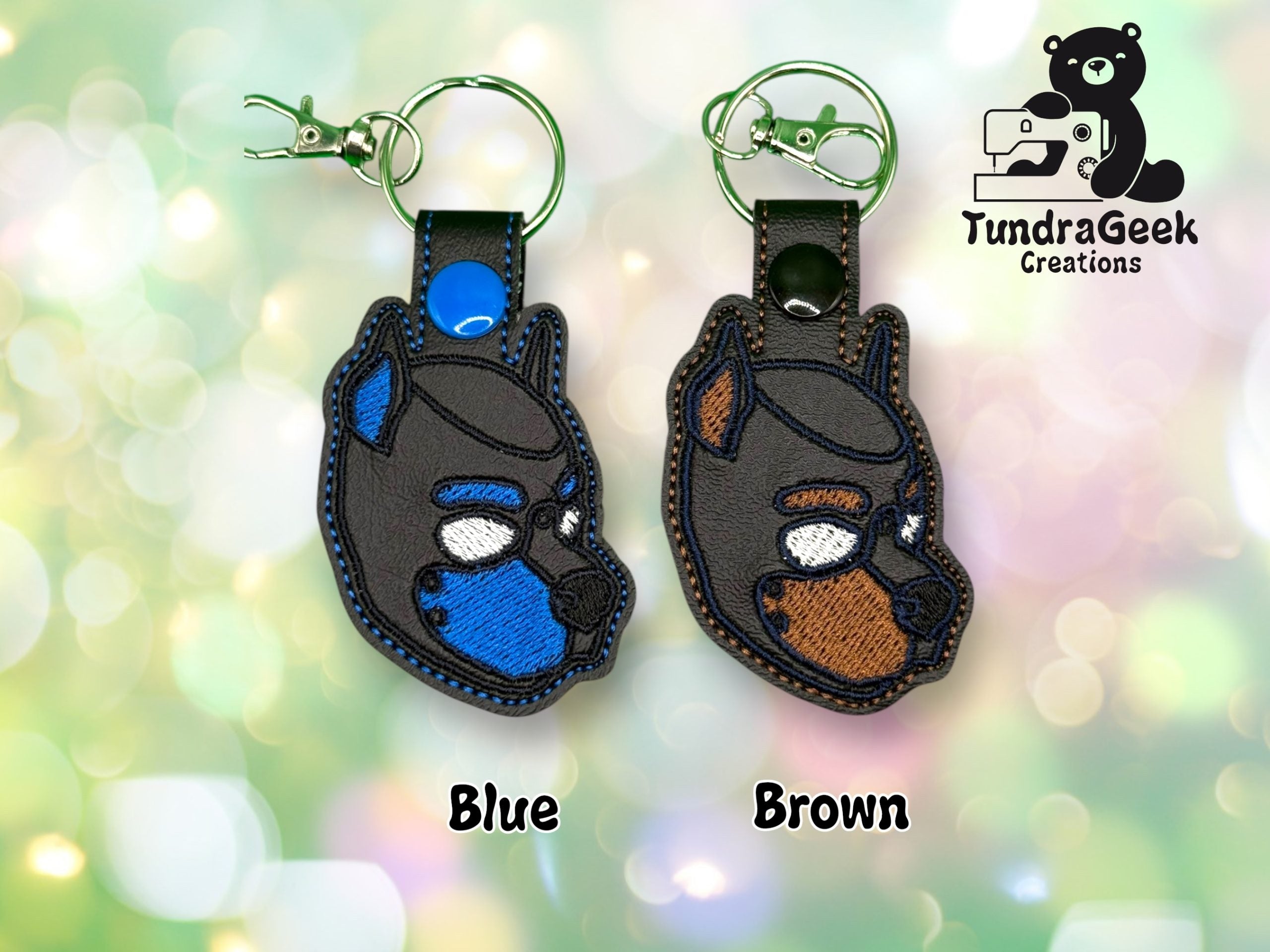 Two embroidered keychains shaped like dog heads, one with blue accents and one with brown accents, displayed on a blurred, colorful background. TundraGeek Creations logo is in the corner.