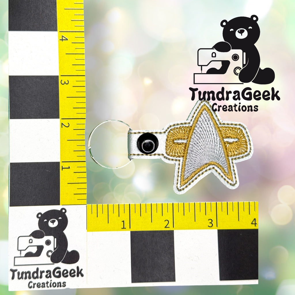 Star Trek-inspired embroidered keychain with a gold and silver insignia, placed on a checkered measurement grid with TundraGeek Creations logo in the background.