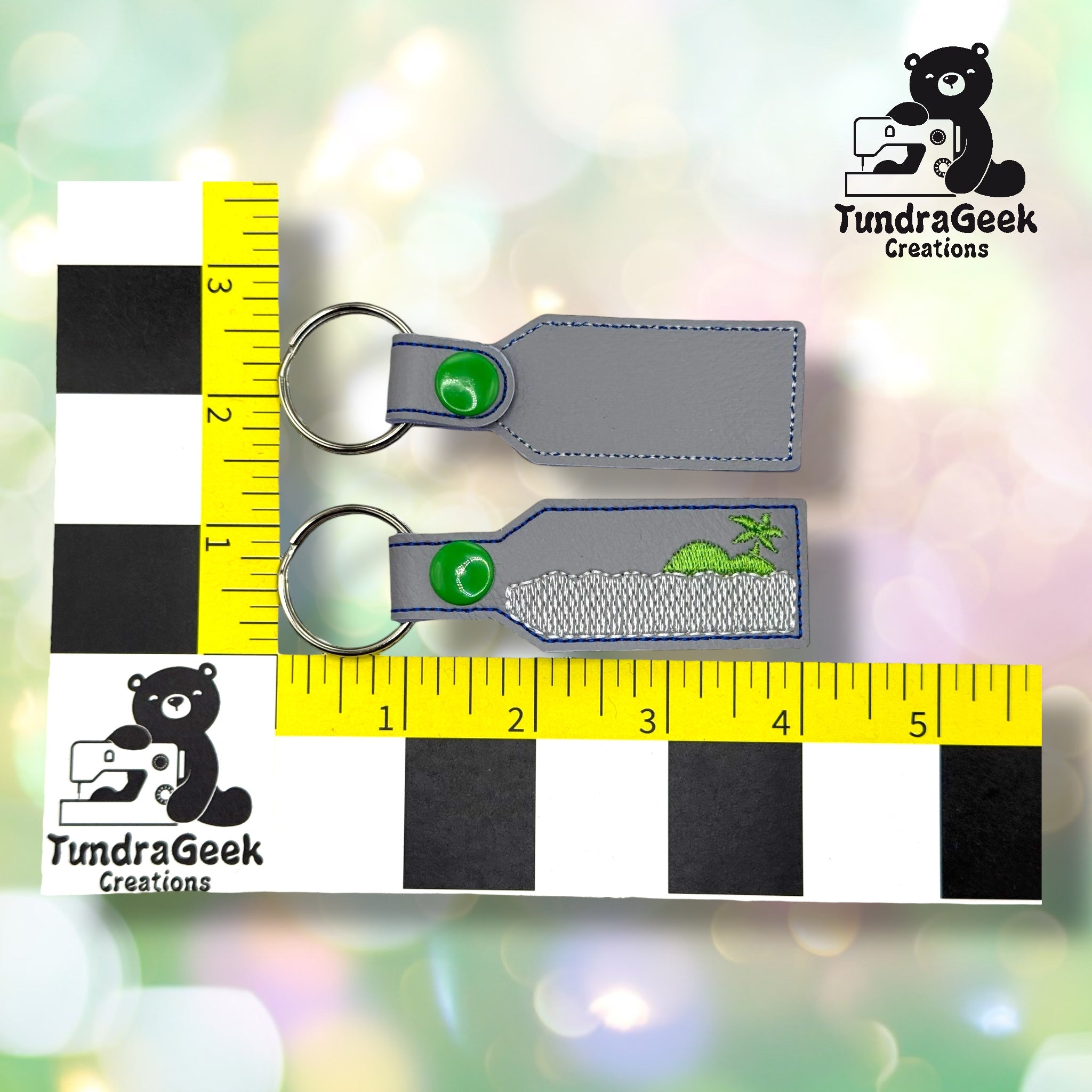 Two gray keychains with green snap buttons and embroidered green dinosaurs, placed on a measurement grid next to a TundraGeek Creations logo.