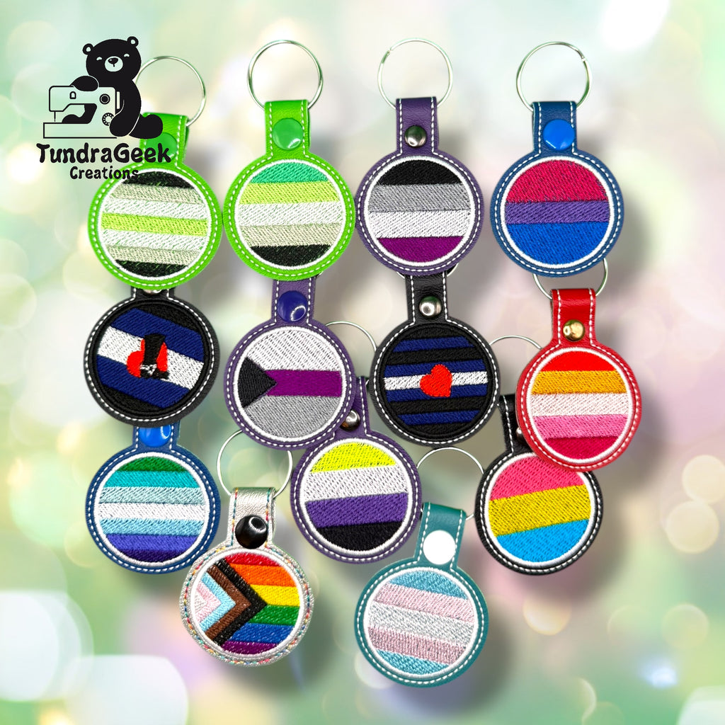 A collection of embroidered keychains featuring various LGBTQIA+ pride flags arranged in rows on a light, bokeh background.
