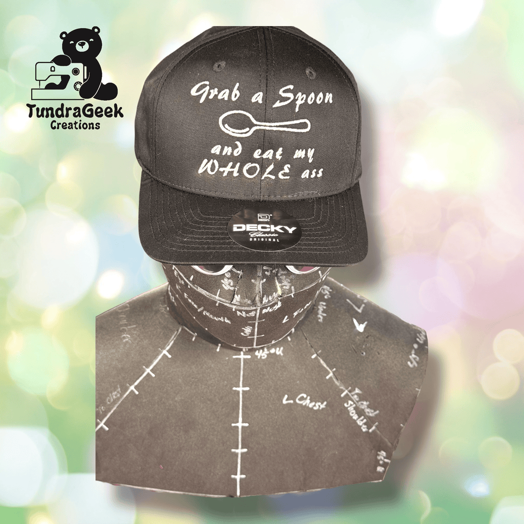 A black baseball cap with the phrase Grab a spoon and eat my whole ass displayed on a mannequin head, with a sewing logo in the top left corner.
