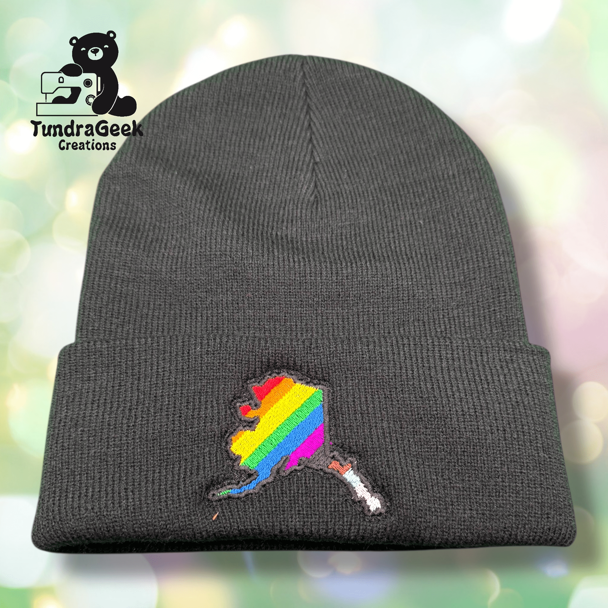 A dark gray knit beanie featuring an embroidered Alaska state outline filled with rainbow pride stripes; TundraGeek Creations logo in the top left corner.