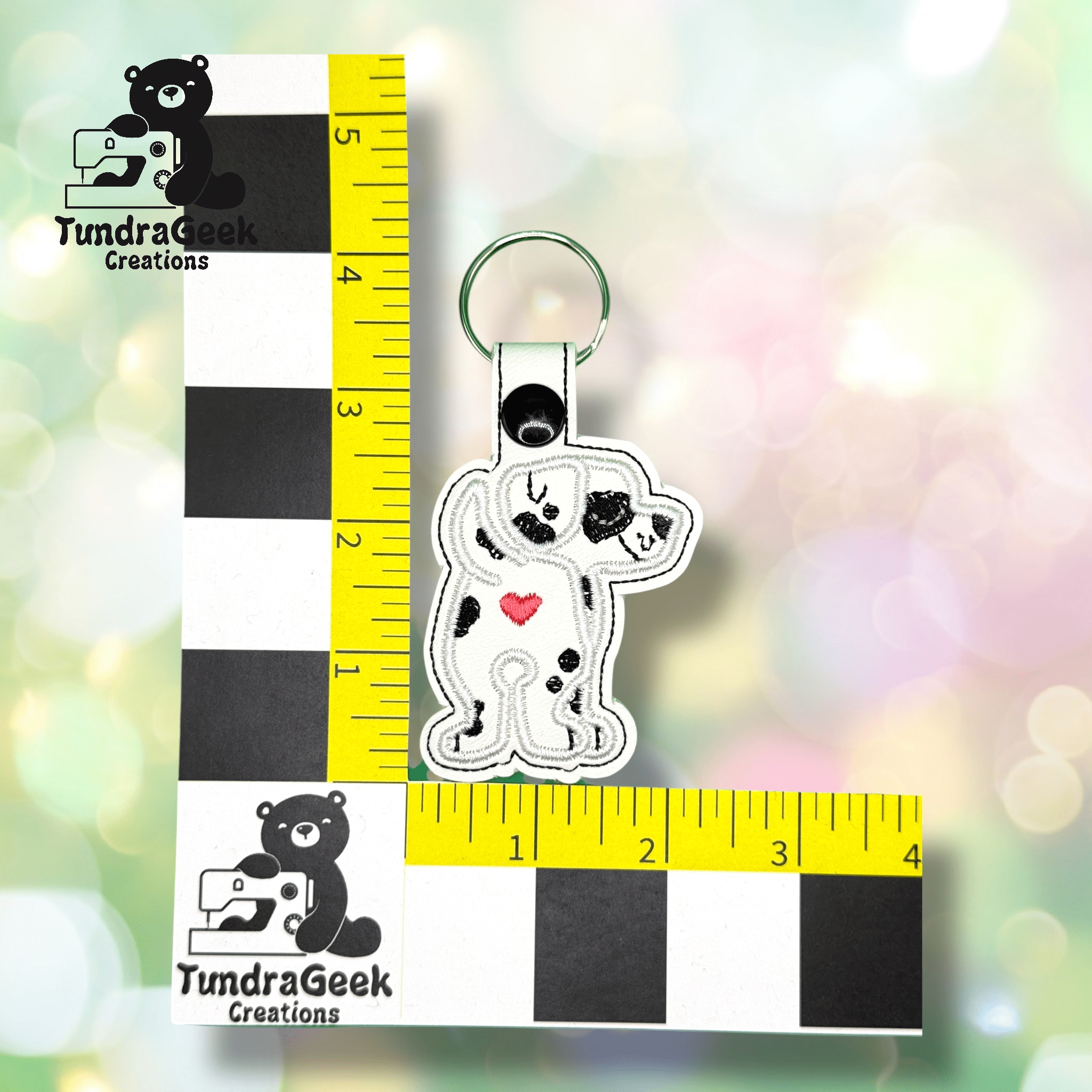 A keychain featuring a white dog with black spots and a red heart, displayed next to a black and yellow measurement grid labeled TundraGeek Creations.