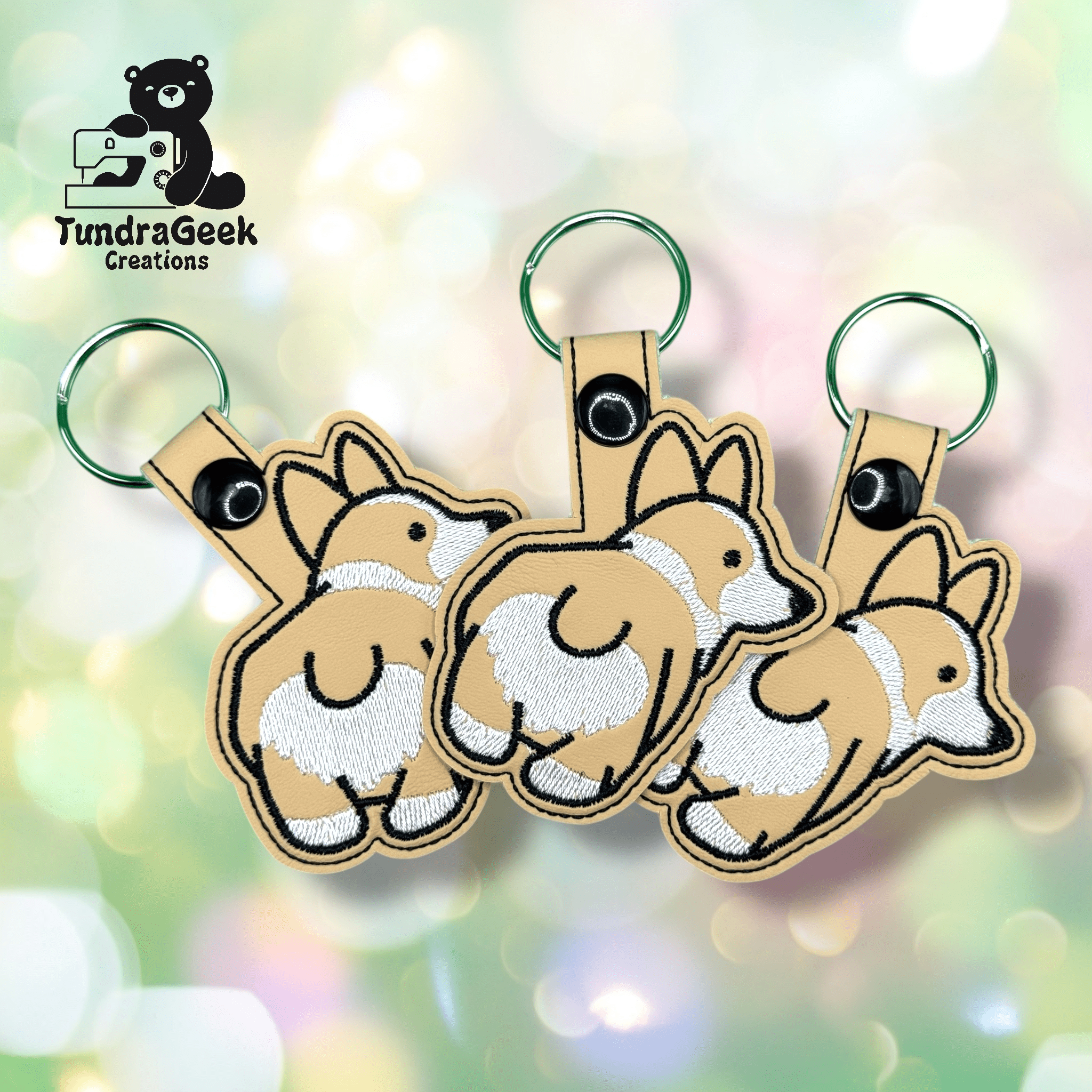 Three embroidered keychains shaped like corgi dogs, showing the back view, are arranged on a blurred pastel background. The TundraGeek Creations logo appears in the top left corner.