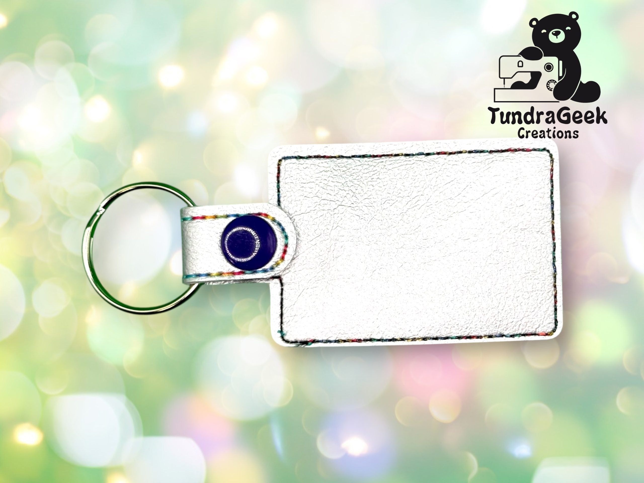 A blank white rectangular keychain with a blue snap and metal ring, displayed on a bokeh background with the TundraGeek Creations logo in the top right corner.