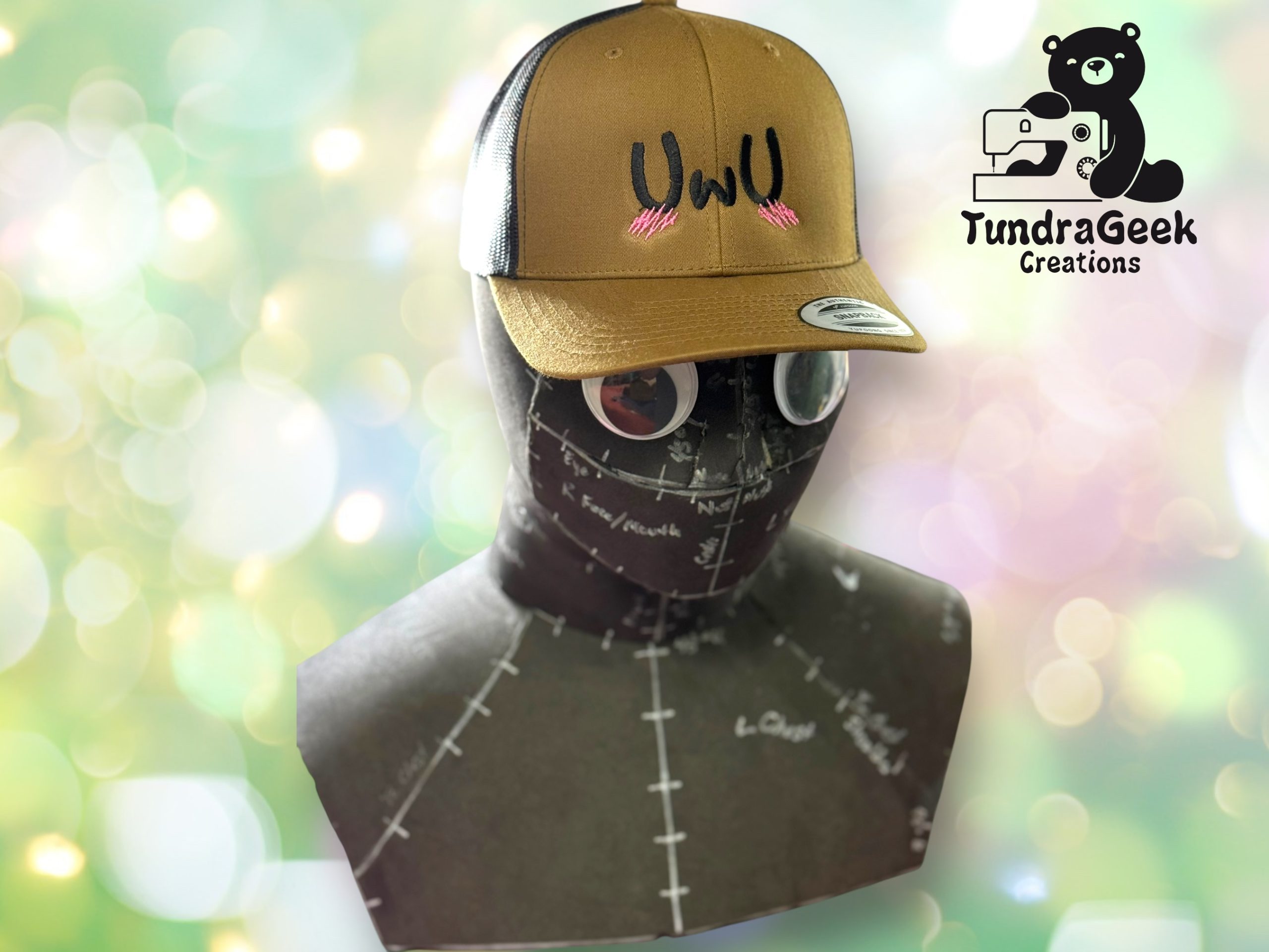A mannequin head wearing a brown and black UWU cap with pink blush marks; TundraGeek Creations logo is in the upper right corner against a blurred background.