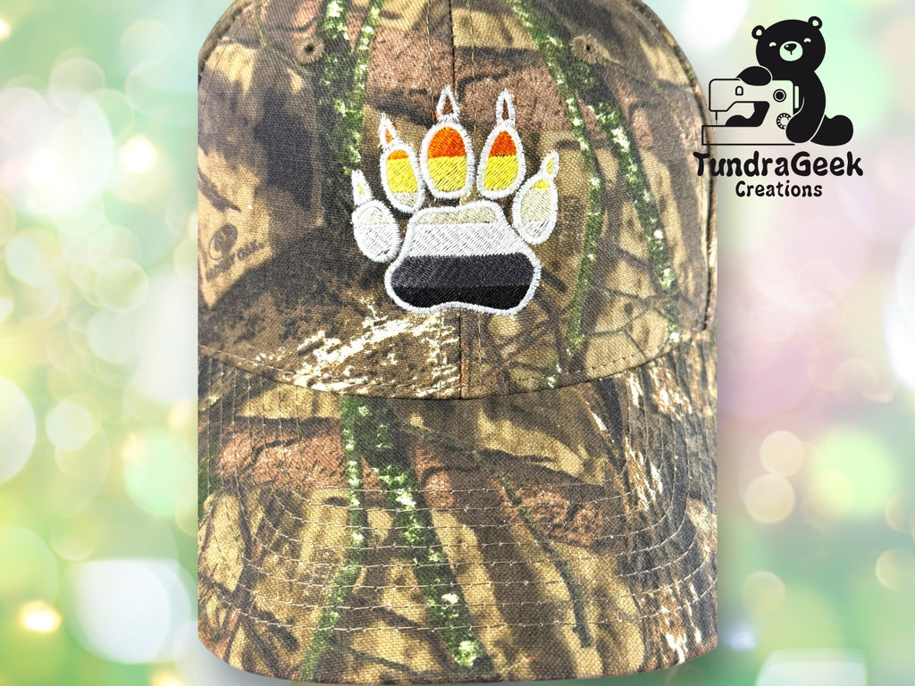 A camouflage baseball cap with a bear paw print in black, gray, white, orange, and yellow, next to the TundraGeek Creations logo on a blurred green background.