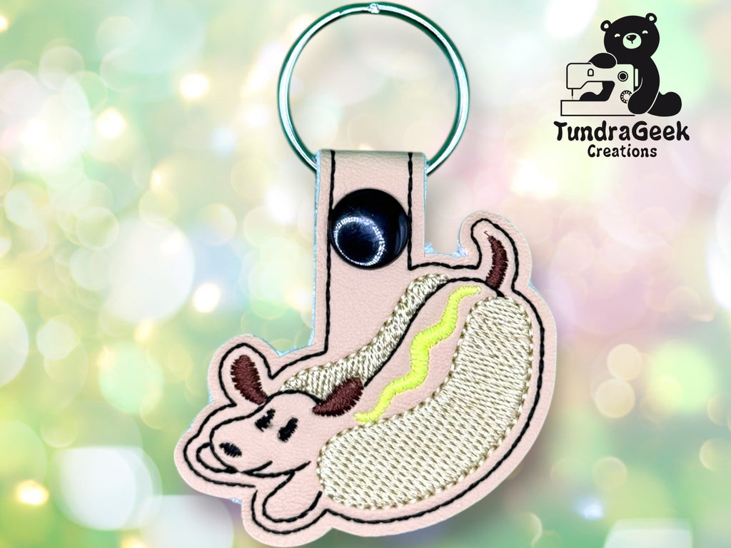 Keychain shaped like a hot dog with a dog’s face embroidered on beige fabric, attached to a metal ring. TundraGeek Creations logo appears in the upper right corner.