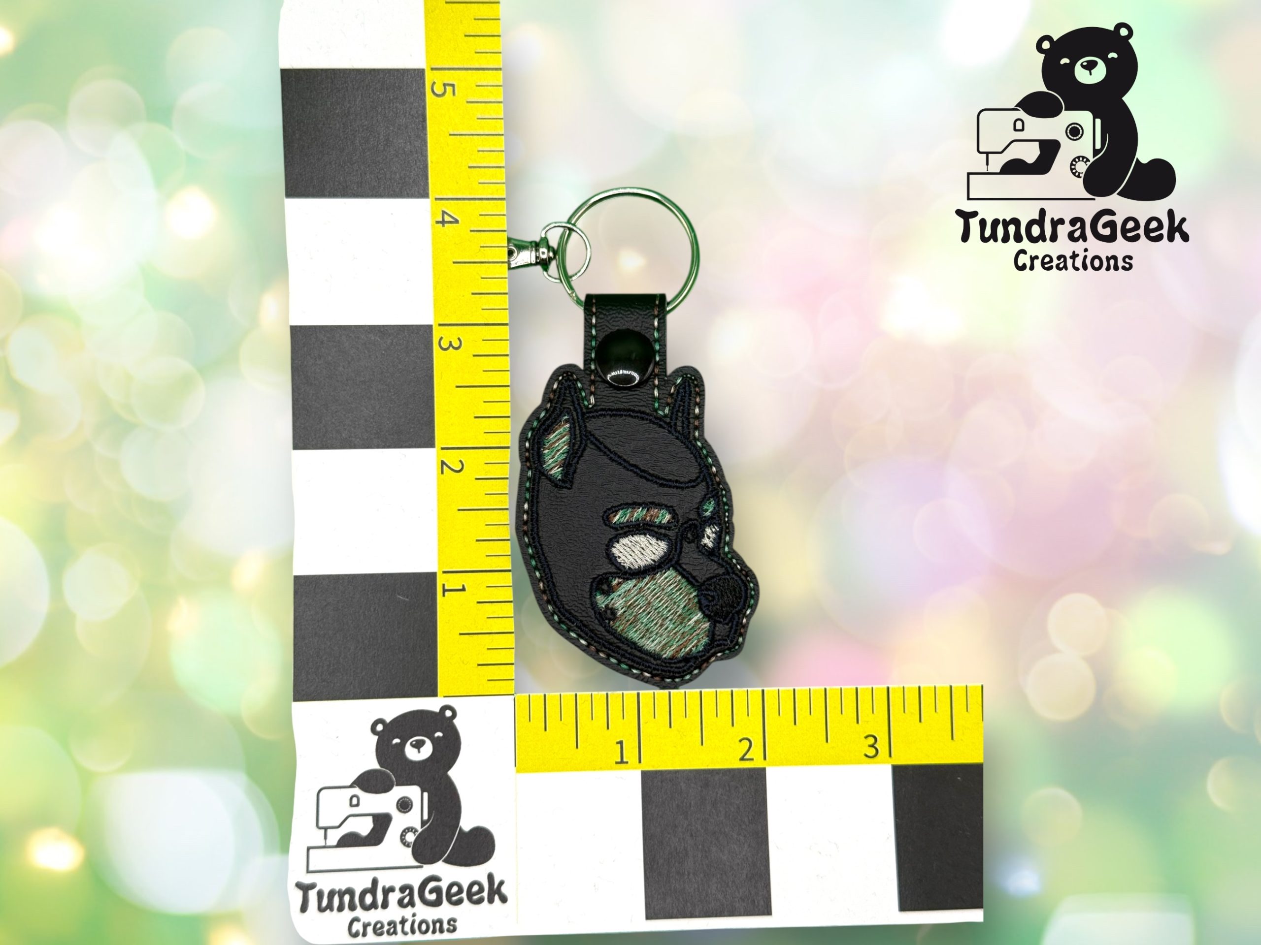 A small keychain shaped like a black bear with green accents is shown next to a ruler for scale. The TundraGeek Creations logo appears in two corners.