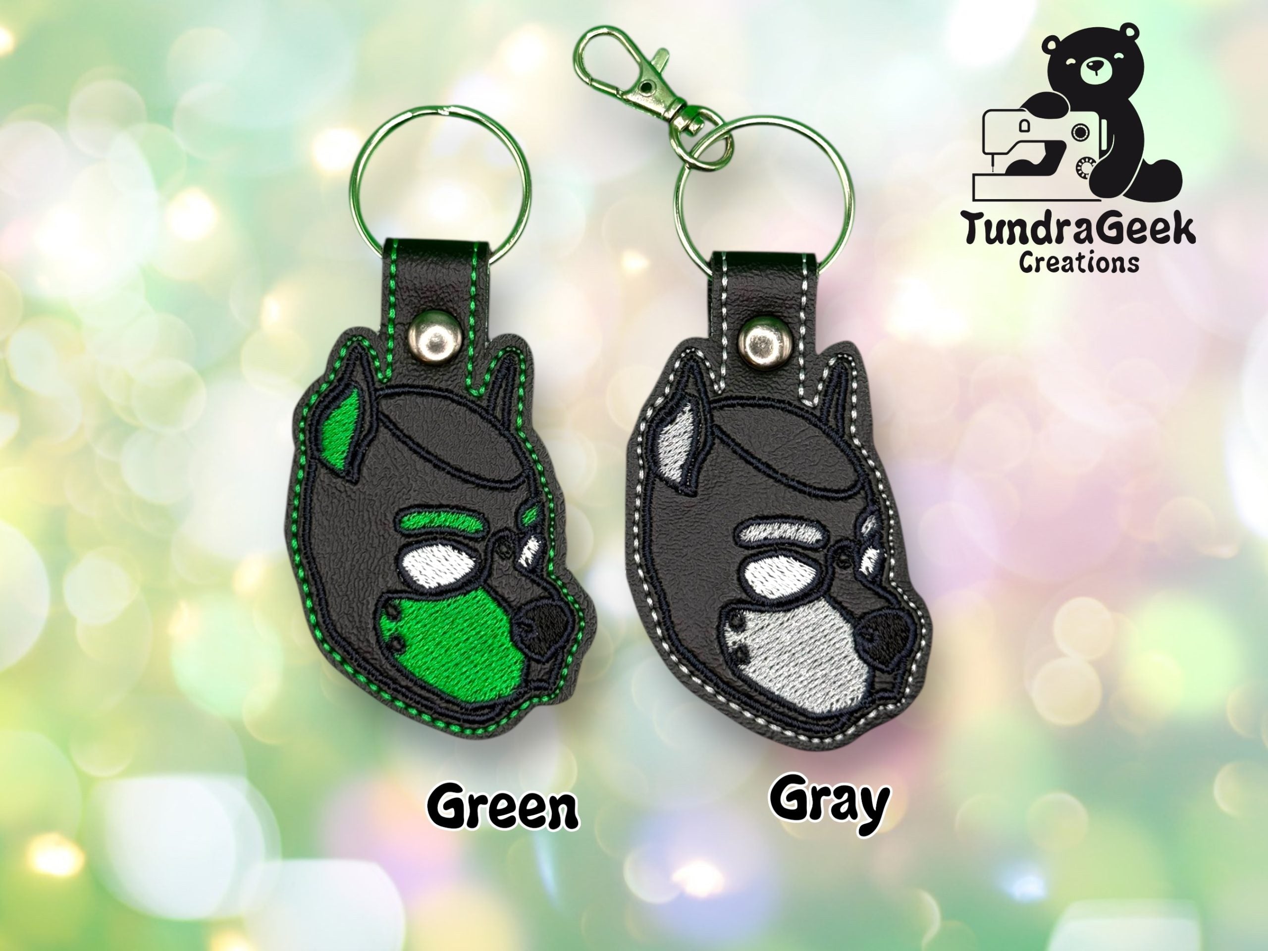Two embroidered keychains shaped like a dogs head, one with green details and one with gray, shown with key rings and a lobster clasp. TundraGeek Creations logo in the top right corner.