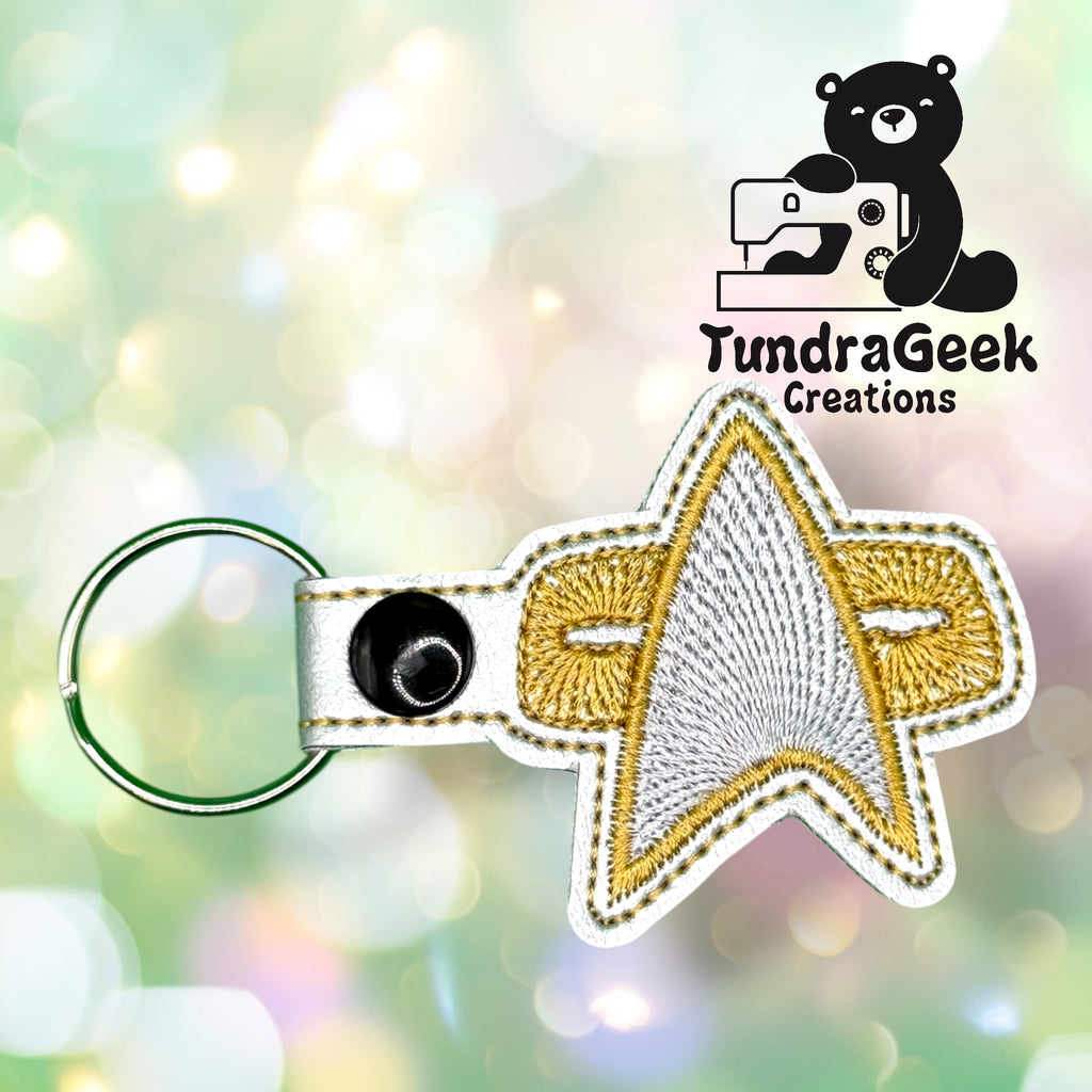 Embroidered keychain in the shape of a gold and silver Star Trek insignia with a snap closure, displayed on a blurred light background. TundraGeek Creations logo in the corner.