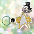 Embroidered gold and silver insignia keychain on a white snap tab. TundraGeek Creations logo in the background.