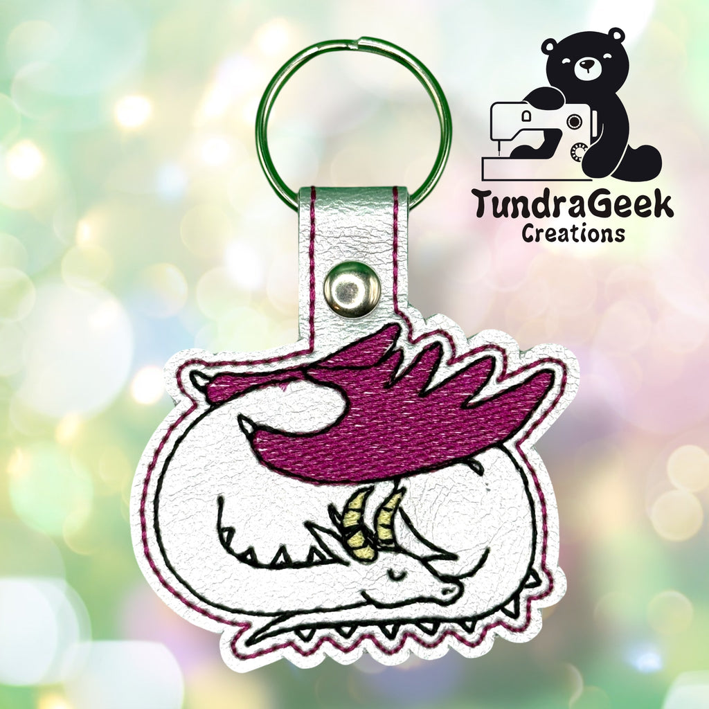 Embroidered keychain featuring a curled sleeping dragon with purple wings and yellow horns, displayed on a blurred light background with the TundraGeek Creations logo.