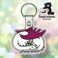Embroidered keychain featuring a curled sleeping dragon with purple wings and yellow horns, displayed on a blurred light background with the TundraGeek Creations logo.