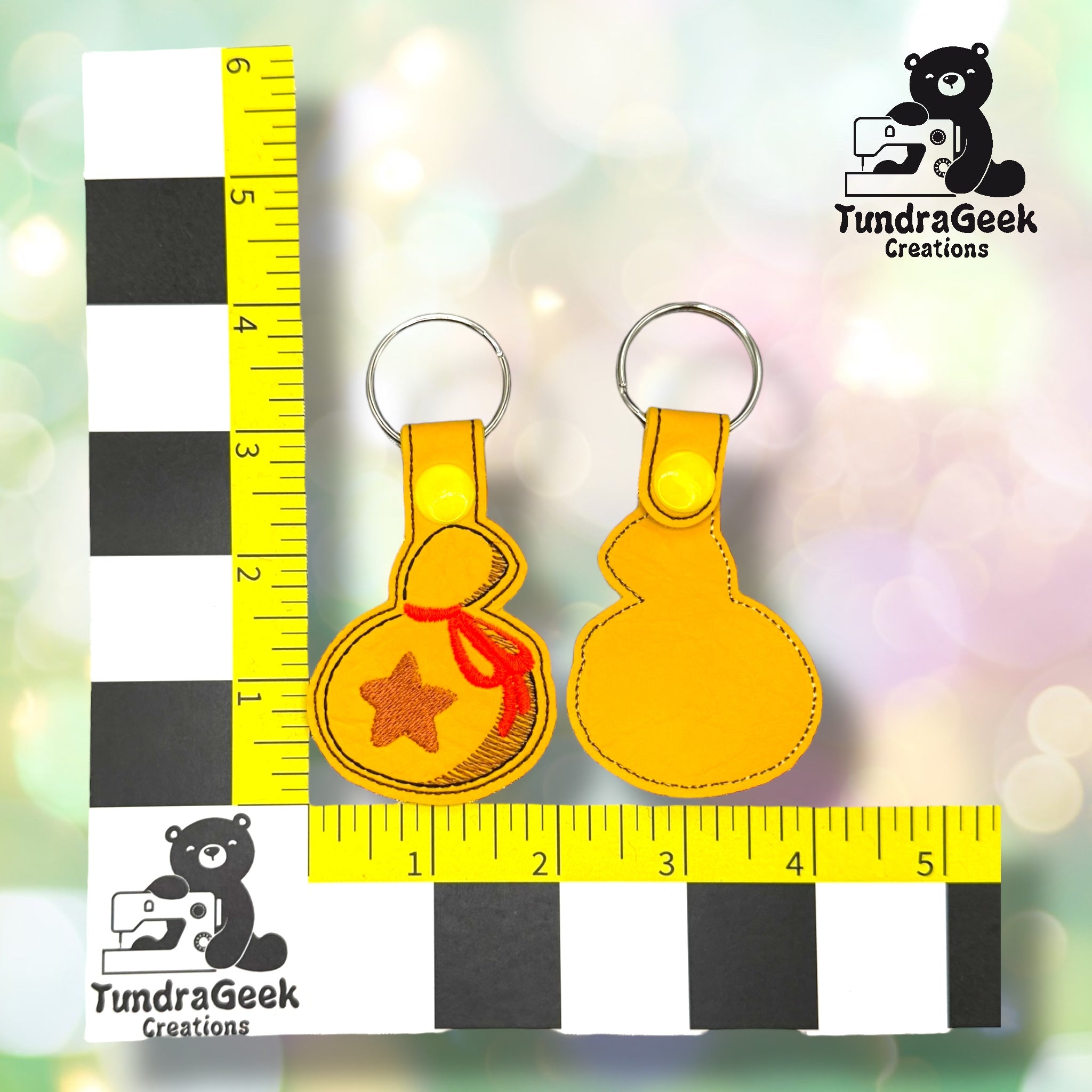 Two yellow keychains shaped like drawstring bags, one with a red star design, are displayed next to a ruler for scale showing the TundraGeek Creations logo.