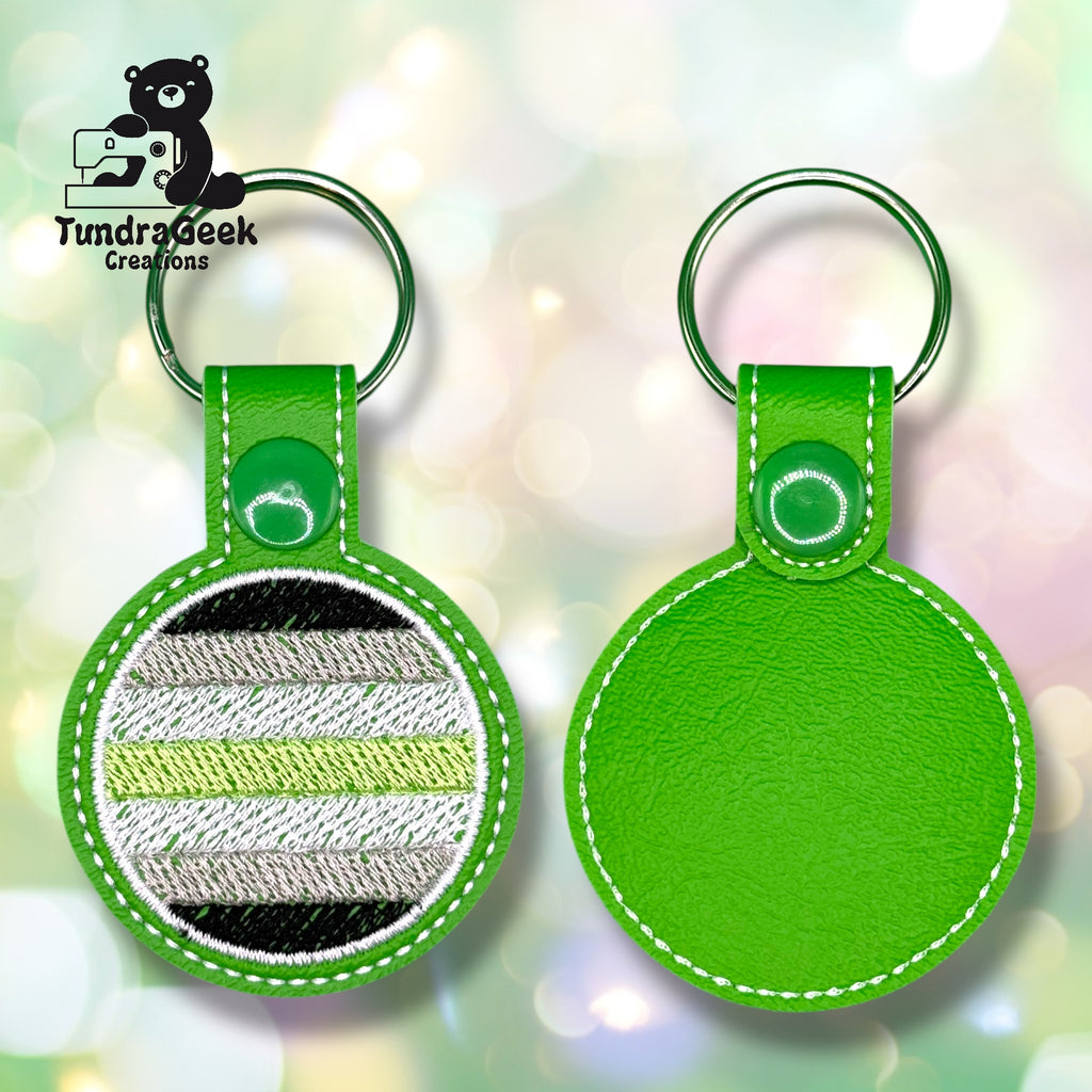 Two round green leather keychains; one shows a striped black, gray, white, and green flag pattern, while the other displays a plain green back. Logo in upper left corner.