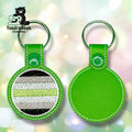 Two round green leather keychains; one shows a striped black, gray, white, and green flag pattern, while the other displays a plain green back. Logo in upper left corner.