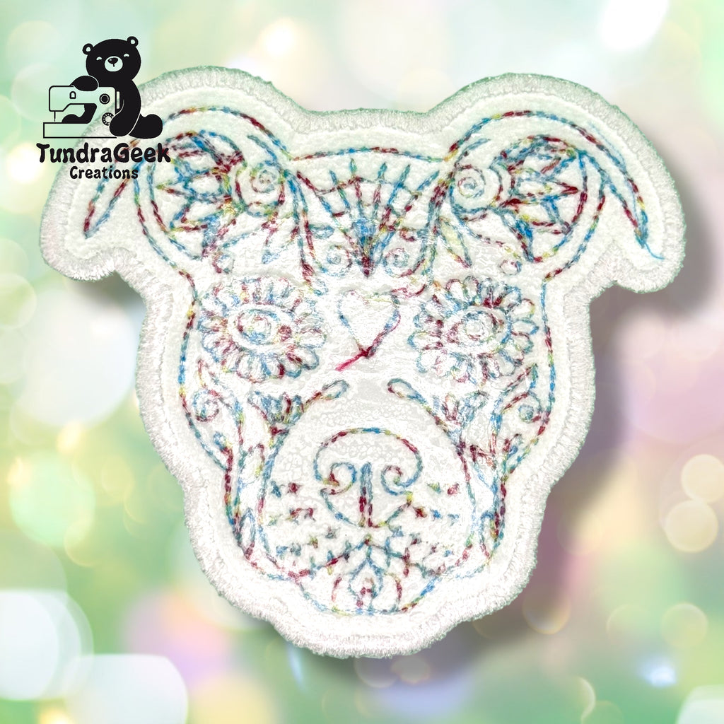 Embroidered patch of a dogs face with colorful thread, featuring floral and geometric patterns, on a blurred light background. TundraGeek Creations logo in the top left corner.