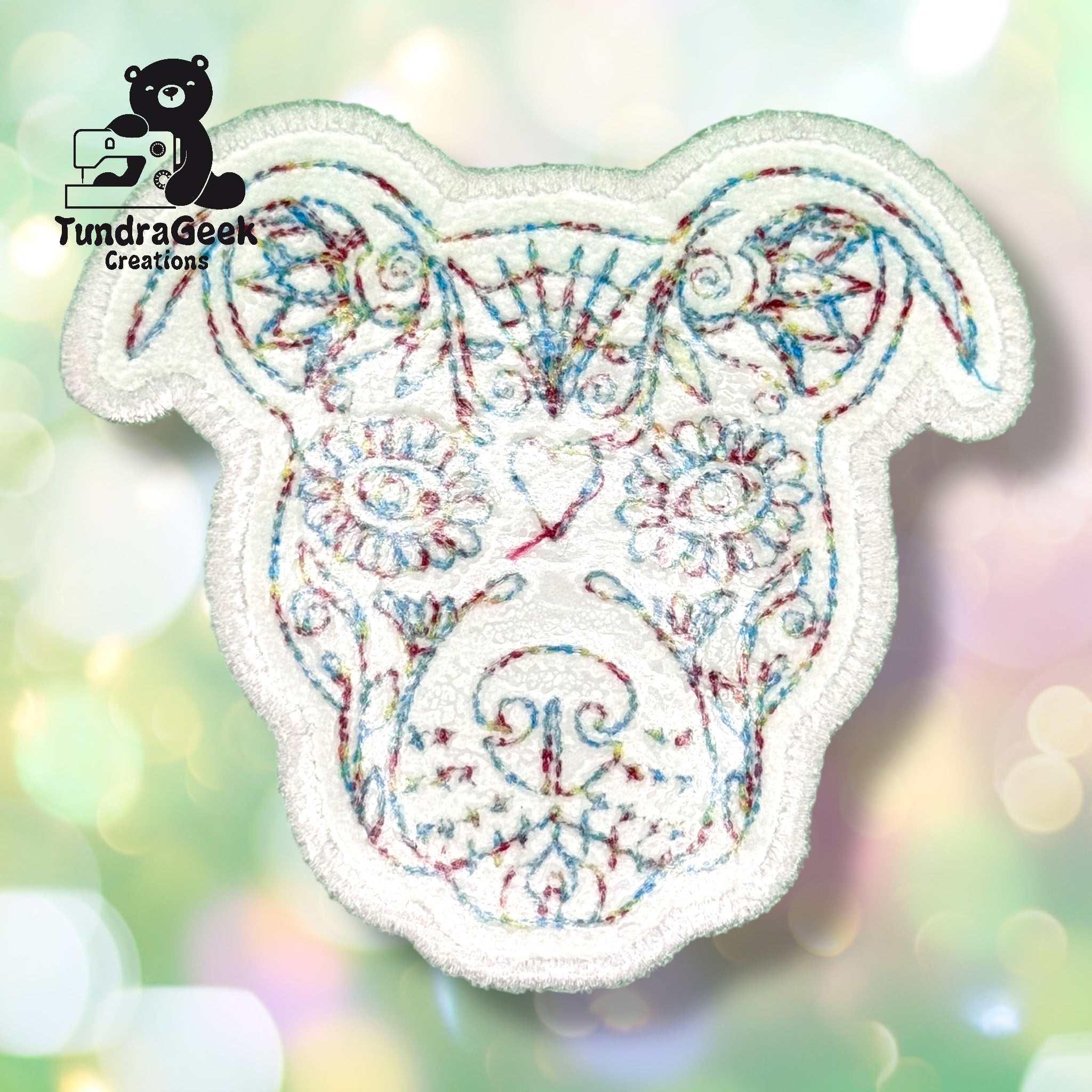 Embroidered patch of a dogs face with colorful thread, featuring floral and geometric patterns, on a blurred light background. TundraGeek Creations logo in the top left corner.