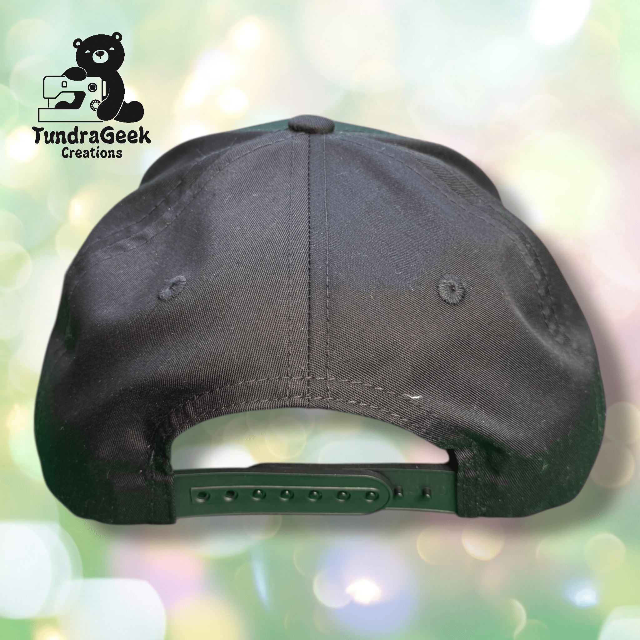 A black snapback hat viewed from the back, displayed against a blurred pastel background with the TundraGeek Creations logo in the top left corner.