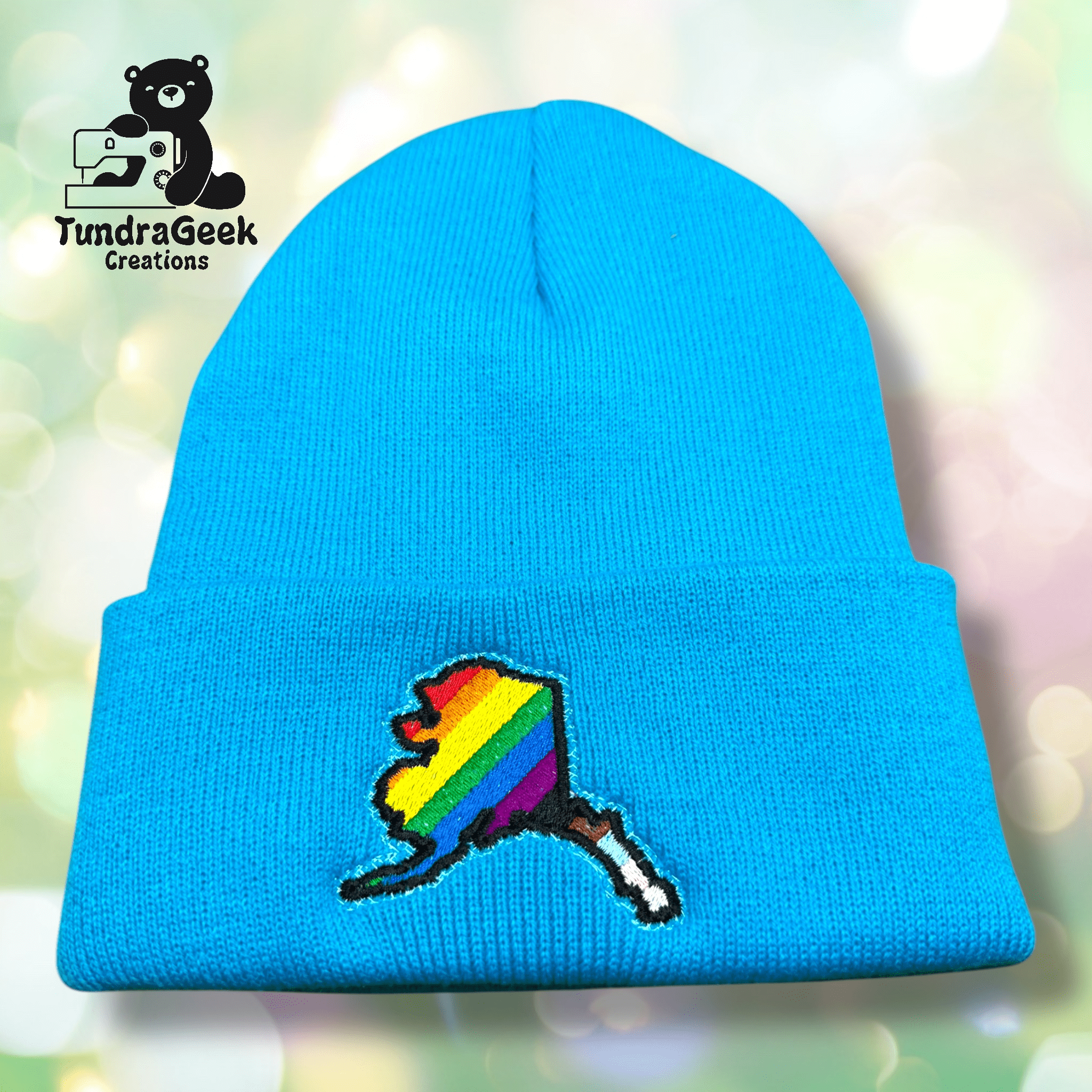 A bright blue knit beanie with an embroidered Alaska state outline in rainbow stripes, displayed in front of a soft, blurred background. TundraGeek Creations logo is in the top left corner.