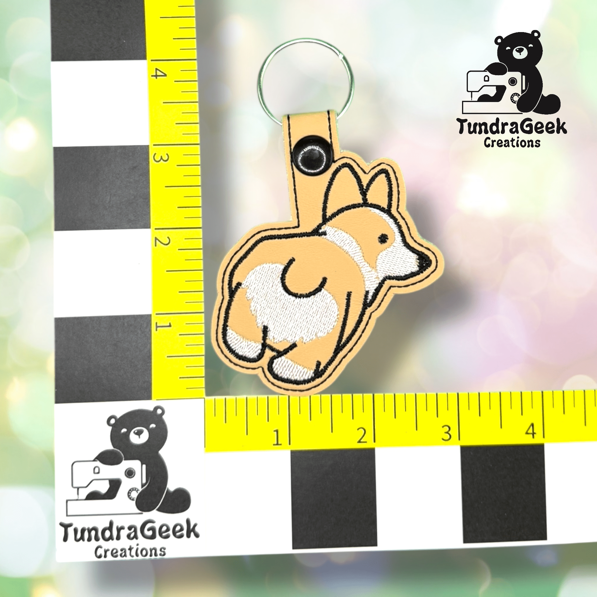 A keychain featuring an embroidered cartoon corgi dog, measured against a yellow and black ruler for scale. The TundraGeek Creations logo appears in the image.