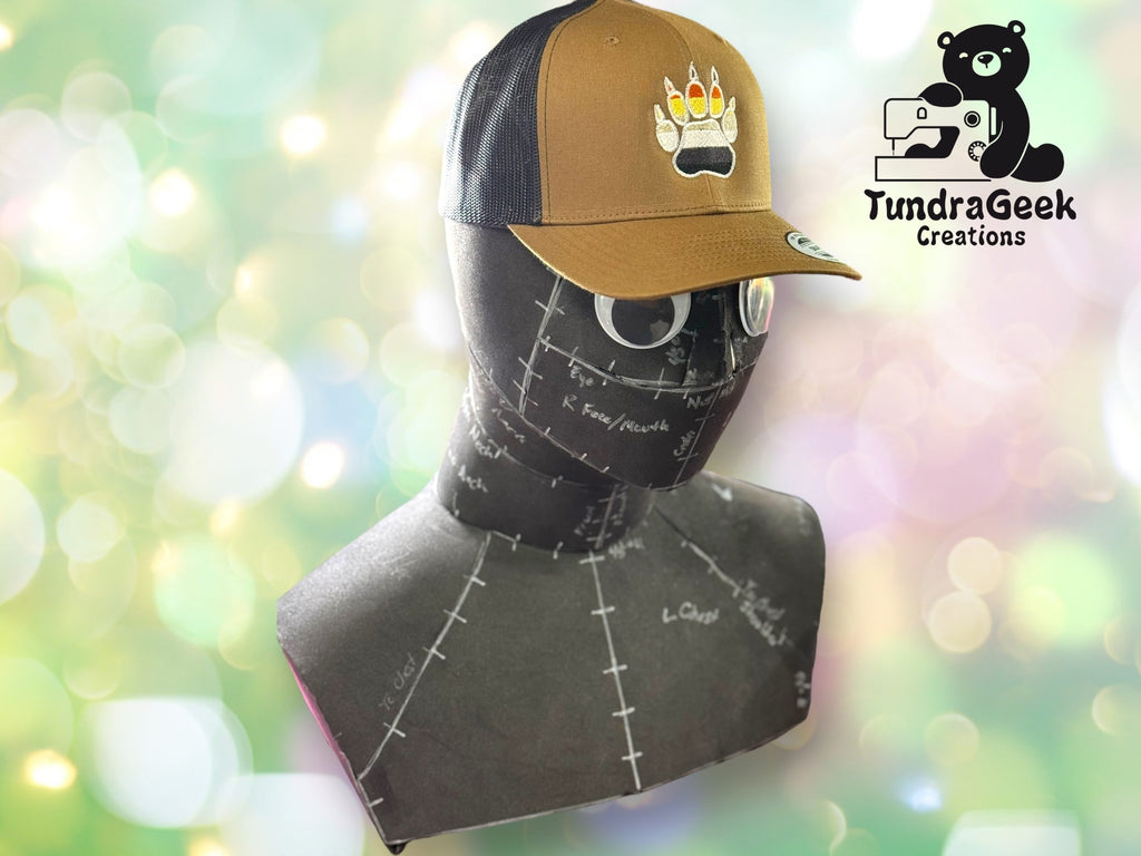 A mannequin head displays a brown and black trucker hat with a bear paw print design; TundraGeek Creations logo is in the upper right corner on a blurred light background.
