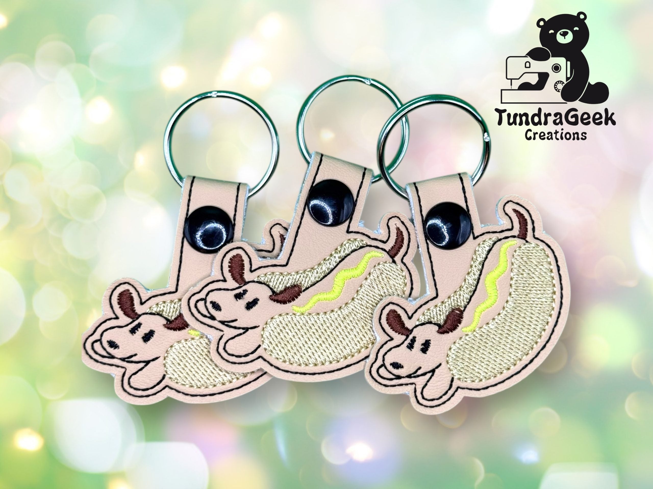Three keychains featuring an embroidered design of a dog in a hot dog bun are shown against a blurred green background with the TundraGeek Creations logo in the upper right.