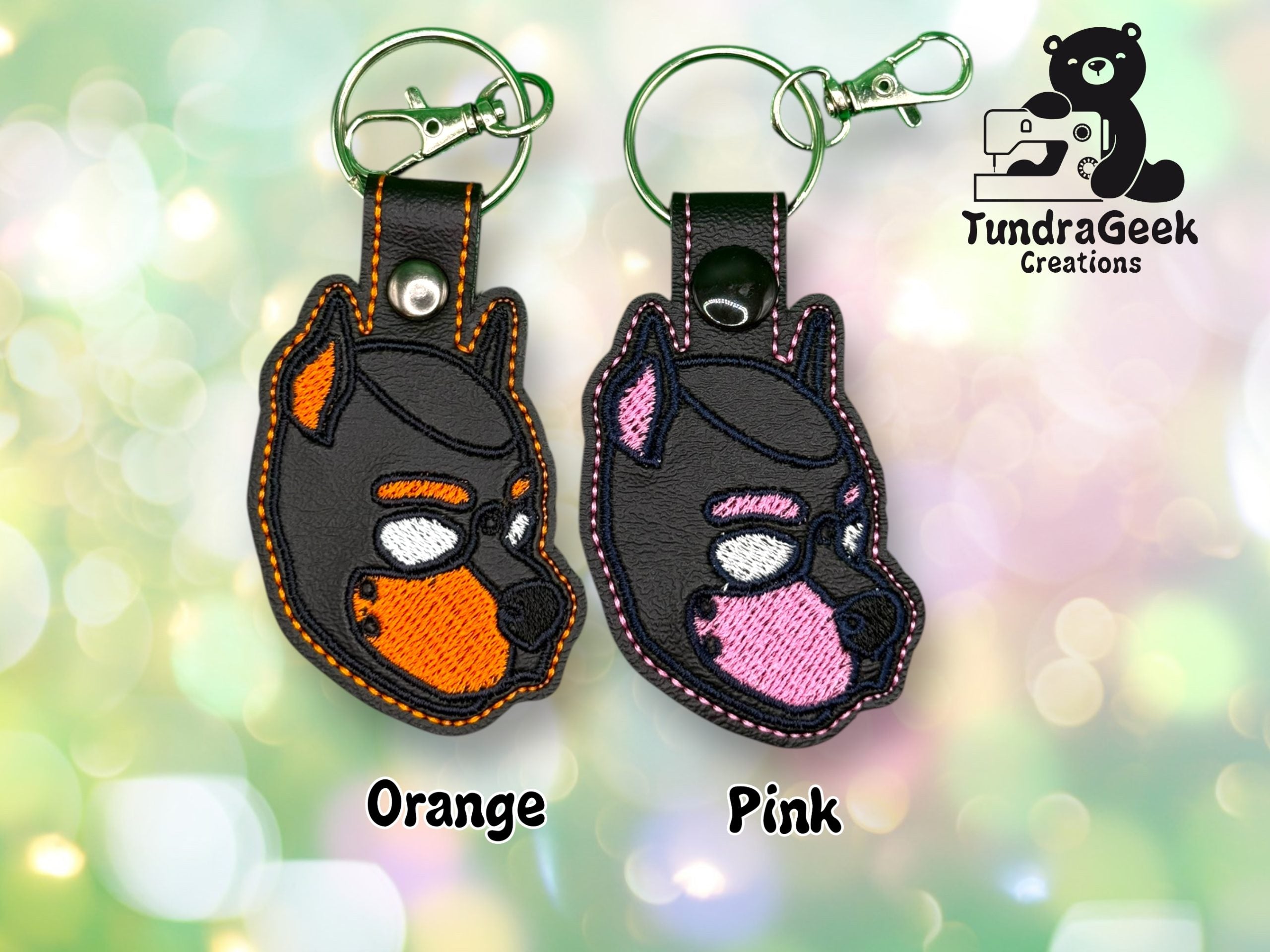 Two embroidered dog face keychains, one with orange accents and one with pink, displayed on a blurred pastel background. TundraGeek Creations logo in the top right corner.
