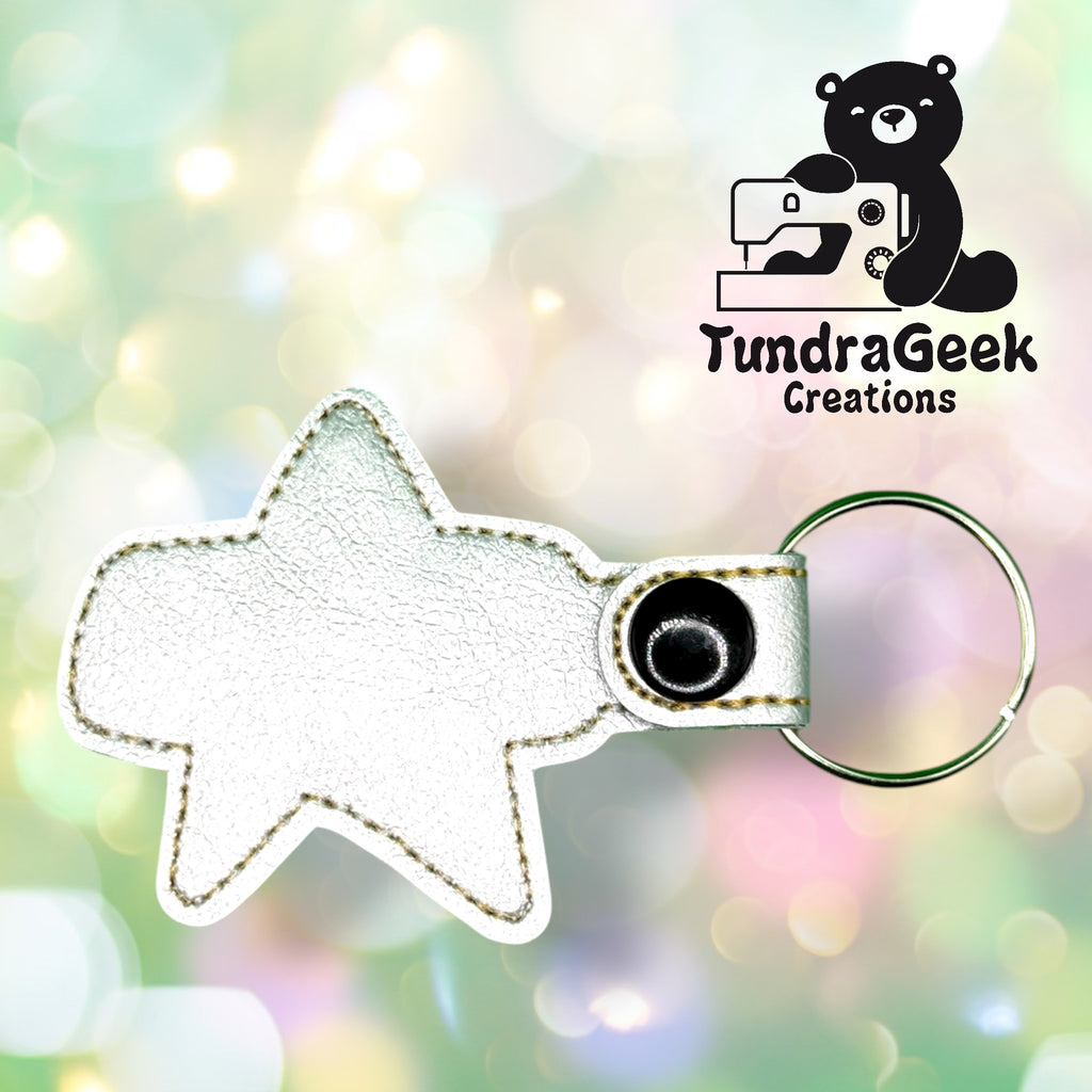 White star-shaped leather keychain with a snap button and key ring, displayed on a pastel bokeh background with the TundraGeek Creations logo in the top right corner.