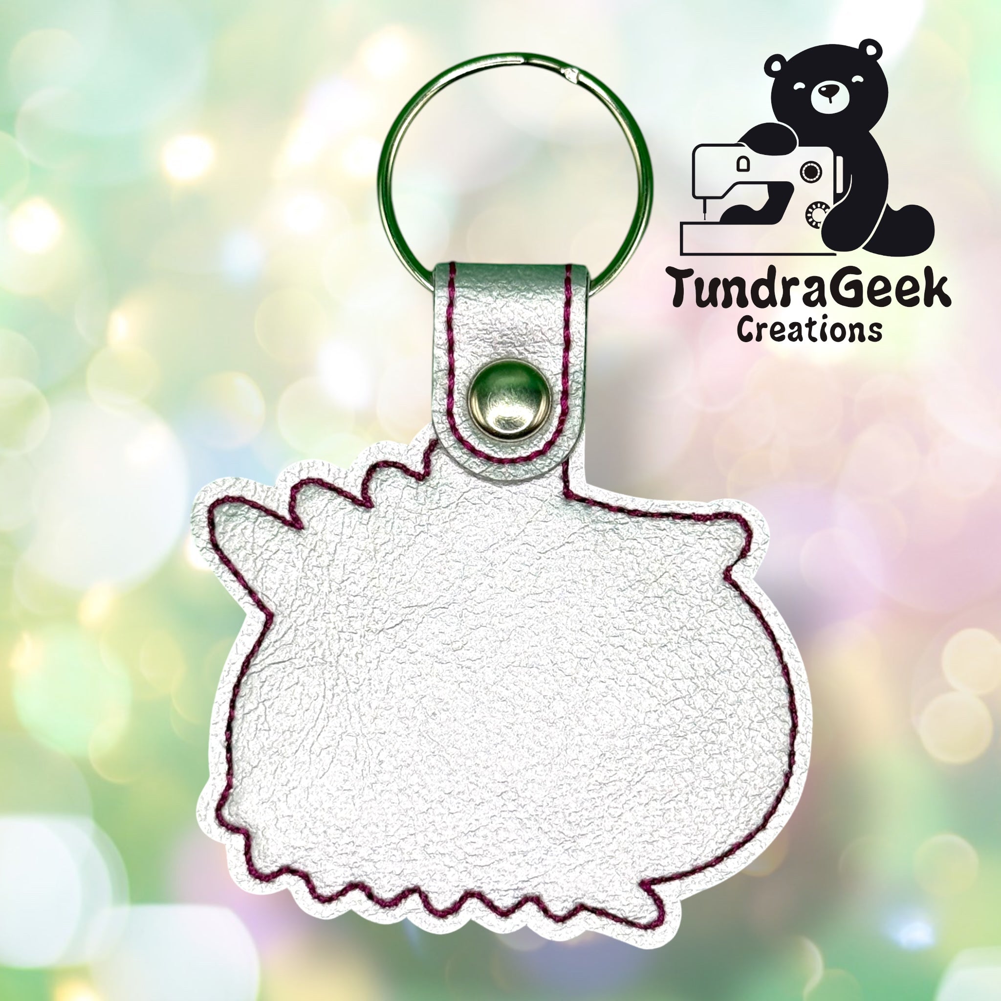 White faux leather keychain with a rounded animal silhouette, outlined in purple stitching, on a blurred bokeh background. TundraGeek Creations logo with a sewing bear is in the top right corner.