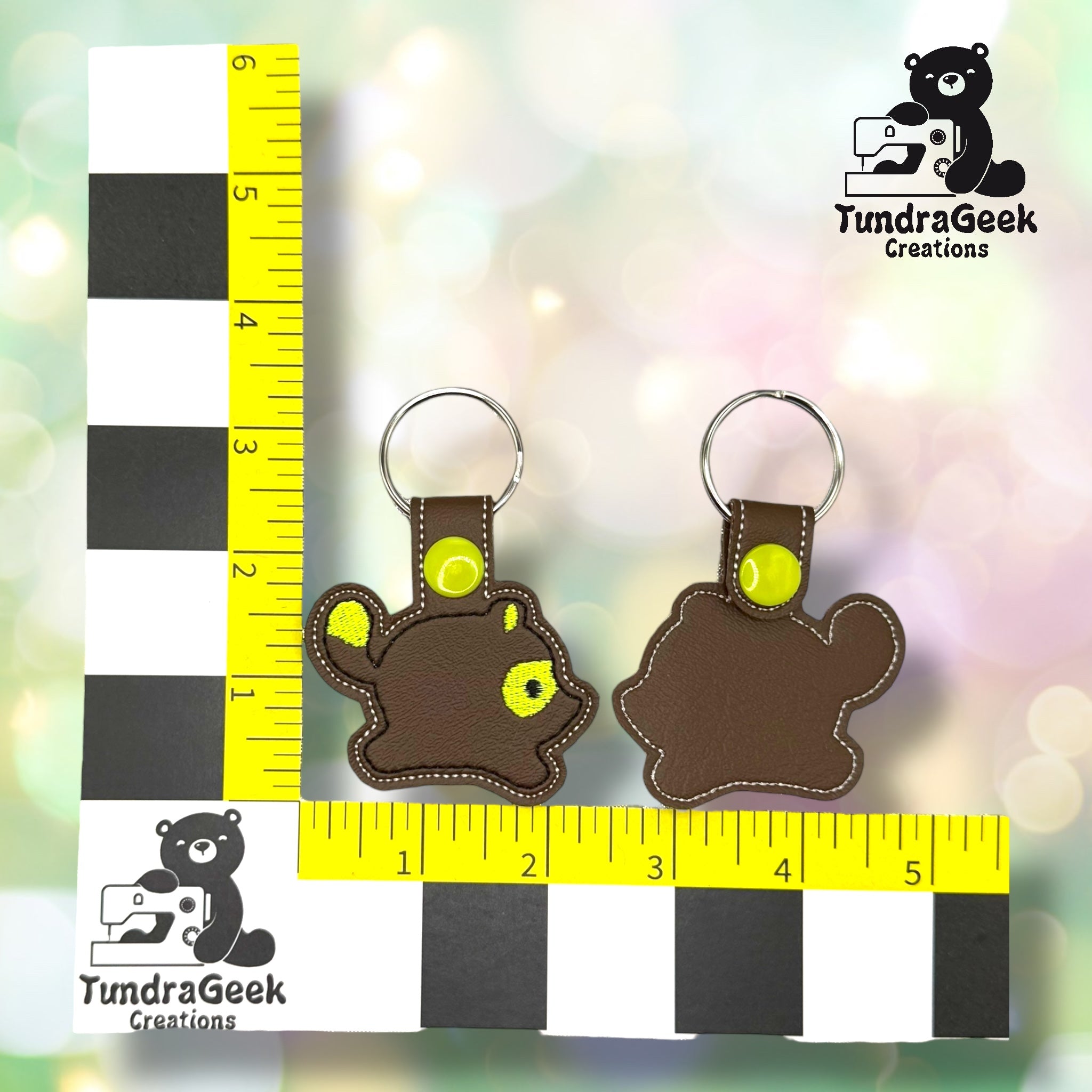 Two brown bear-shaped keychains with yellow accents are displayed next to rulers for scale. The TundraGeek Creations logo is visible in two corners.