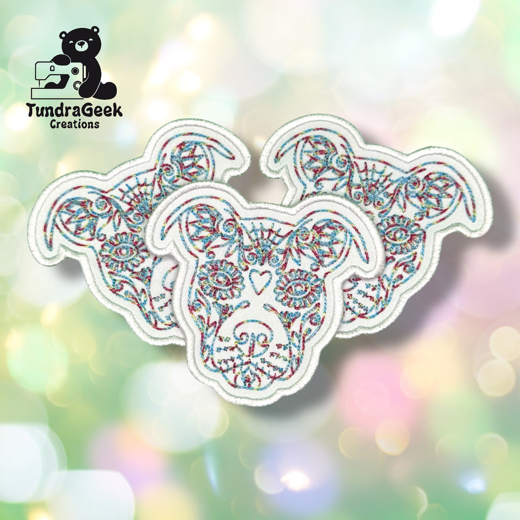 Three identical embroidered dog head patches with intricate multicolored line designs are displayed on a soft, blurred pastel background. TundraGeek Creations logo is in the upper left corner.