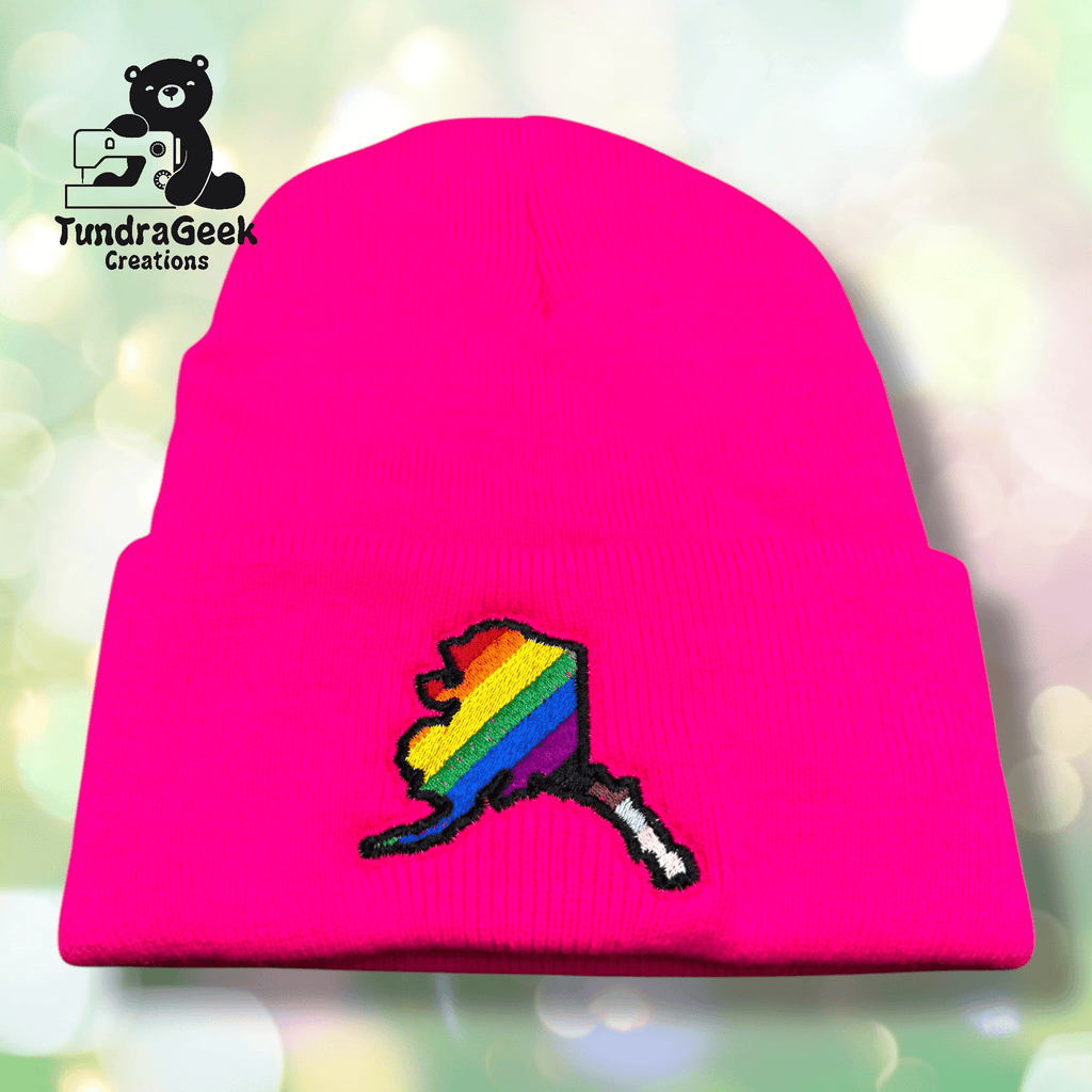 A bright pink beanie featuring an embroidered rainbow pride Michigan state design and the TundraGeek Creations logo in the top left corner.