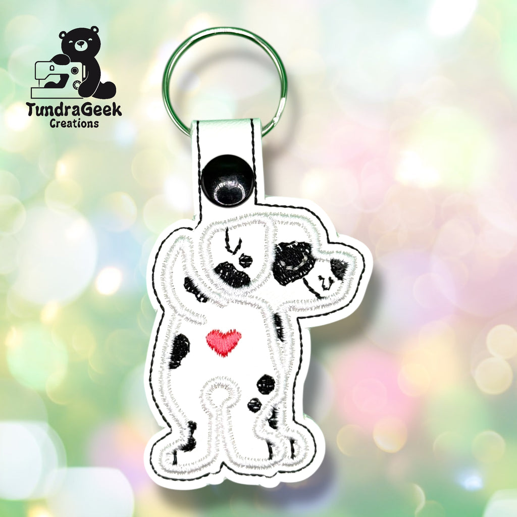 Keychain featuring an embroidered cartoon Dalmatian with black spots and a red heart on its chest, against a blurred pastel background. TundraGeek Creations logo in the top left corner.