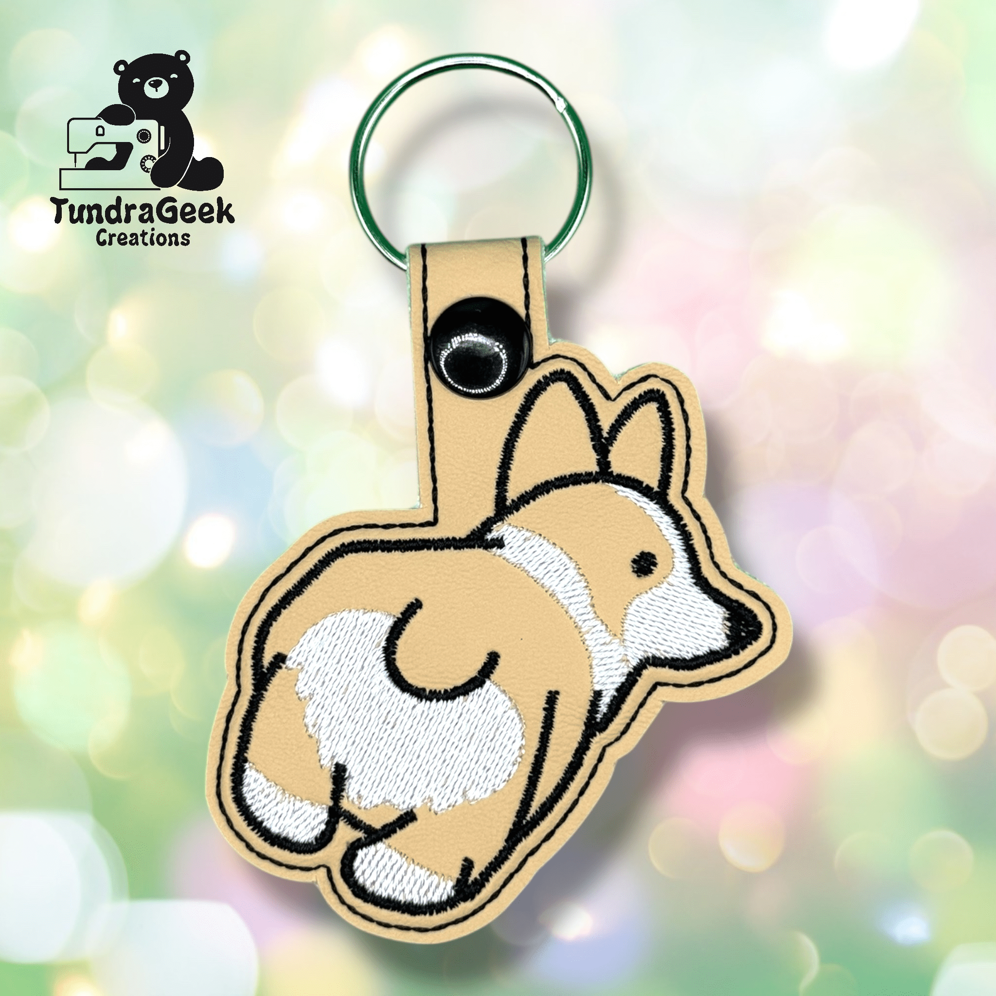 Embroidered keychain shaped like a corgi dog with a visible rear, attached to a green keyring. Logo TundraGeek Creations is in the top left corner.