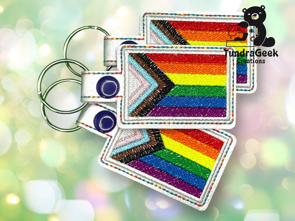 Three keychains with embroidered Progress Pride flags, displayed on a light green bokeh background. TundraGeek Creations logo with a bear appears in the top right corner.