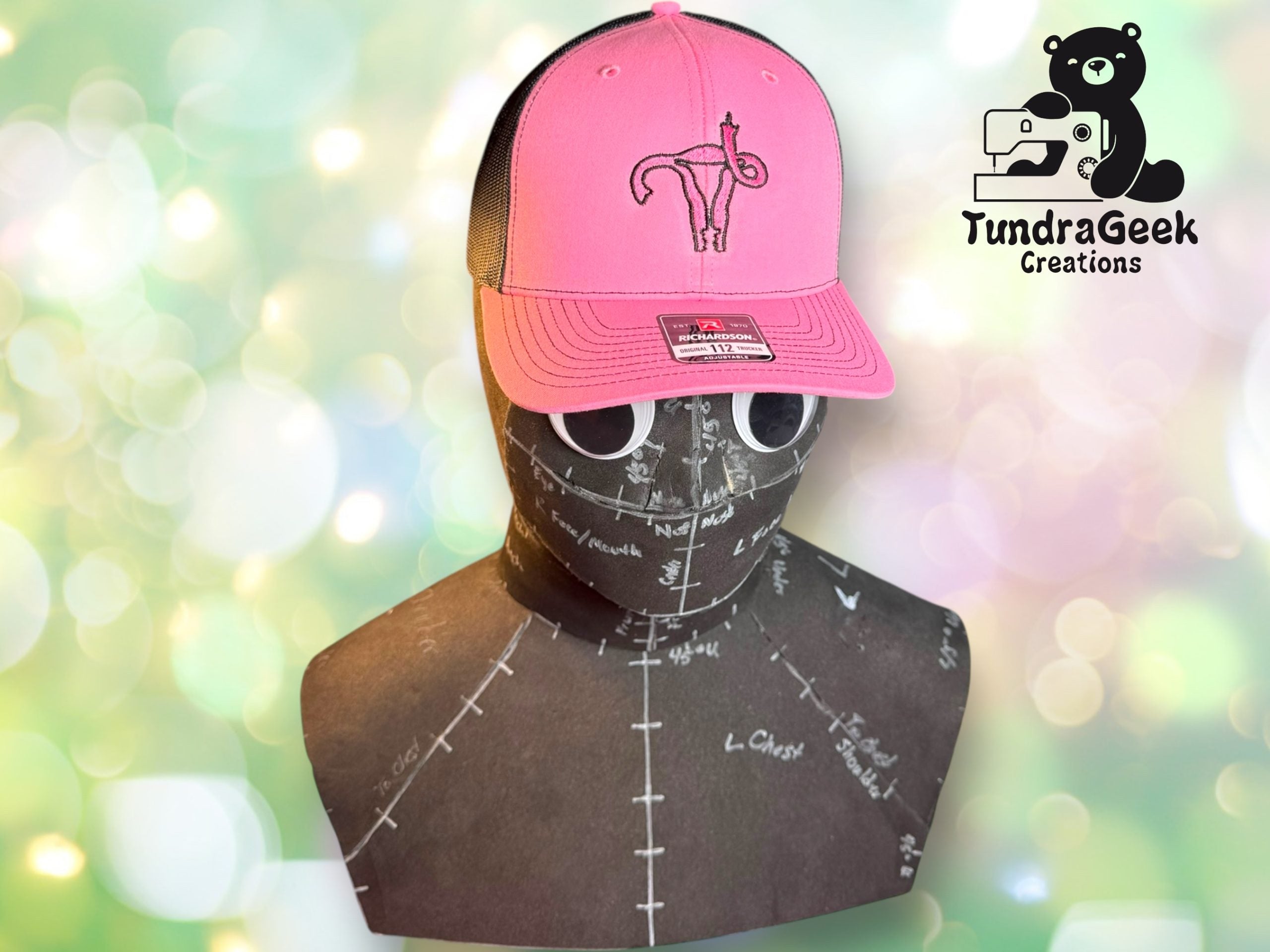 A mannequin head wearing a pink trucker hat with a uterus graphic, in front of a blurred bokeh background and a TundraGeek Creations logo.