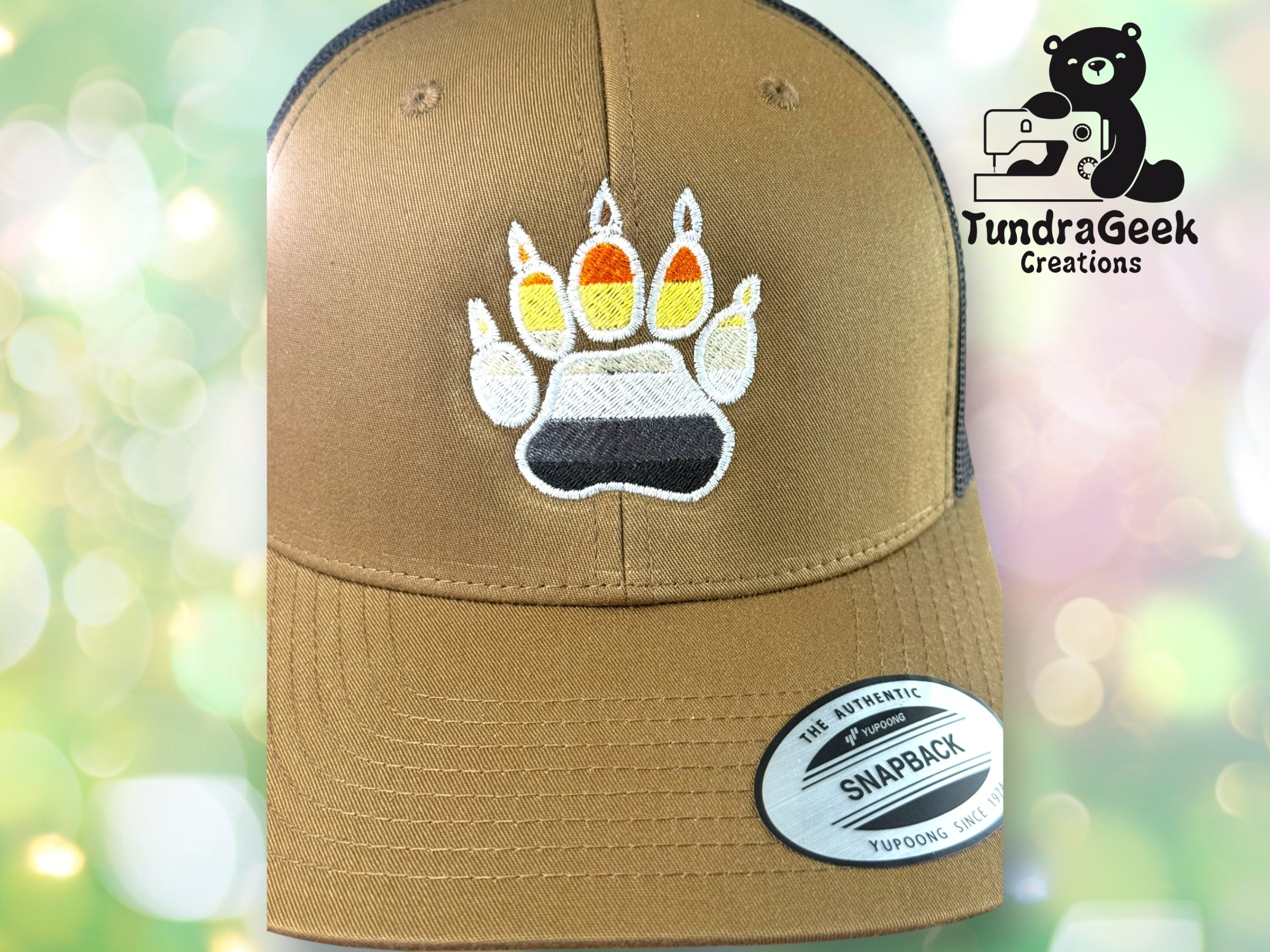 Brown snapback hat featuring an embroidered bear paw print with black, gray, white, yellow, orange, and red colors. TundraGeek Creations logo appears in the upper right corner.