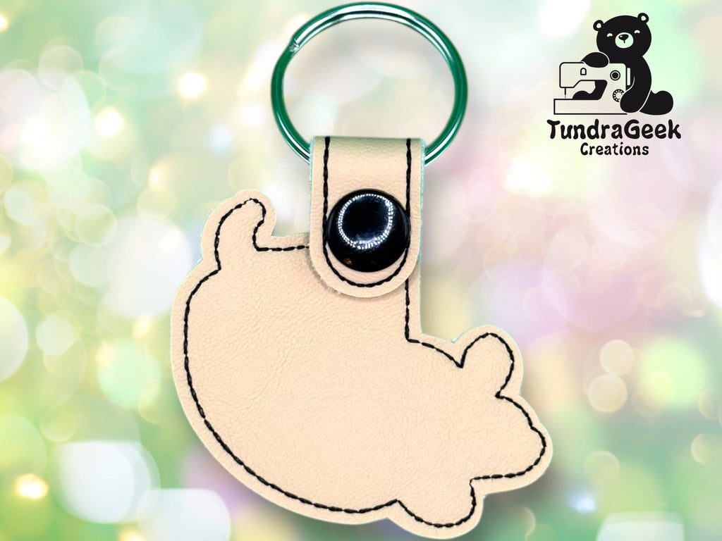 A tan, fish-shaped leather keychain with black stitching, attached to a green metal key ring. The TundraGeek Creations logo with a bear and sewing machine is in the top right corner.