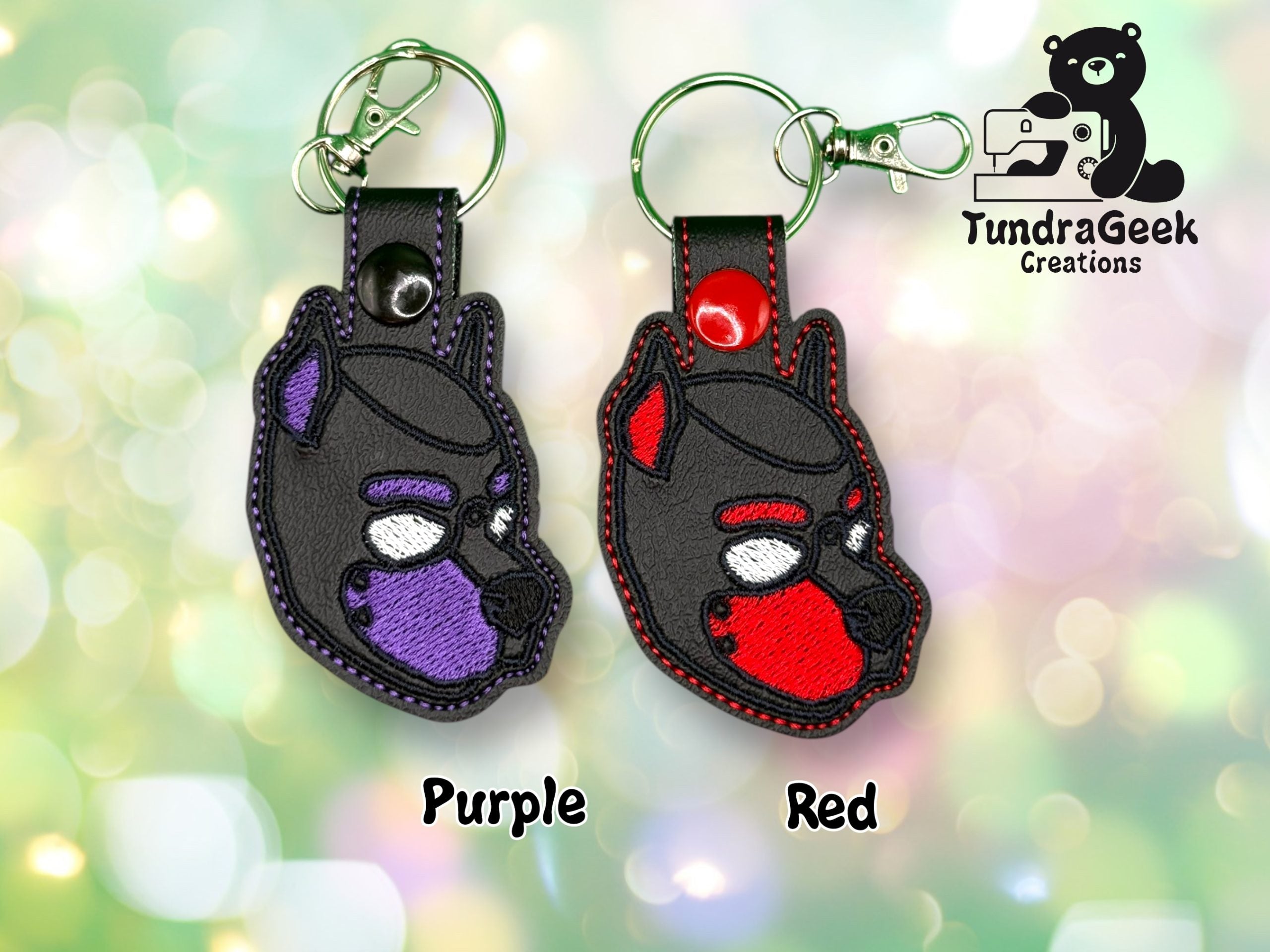 Two embroidered keychains shaped like a dogs head, one with purple accents and one with red accents, displayed on a blurred background with TundraGeek Creations logo.