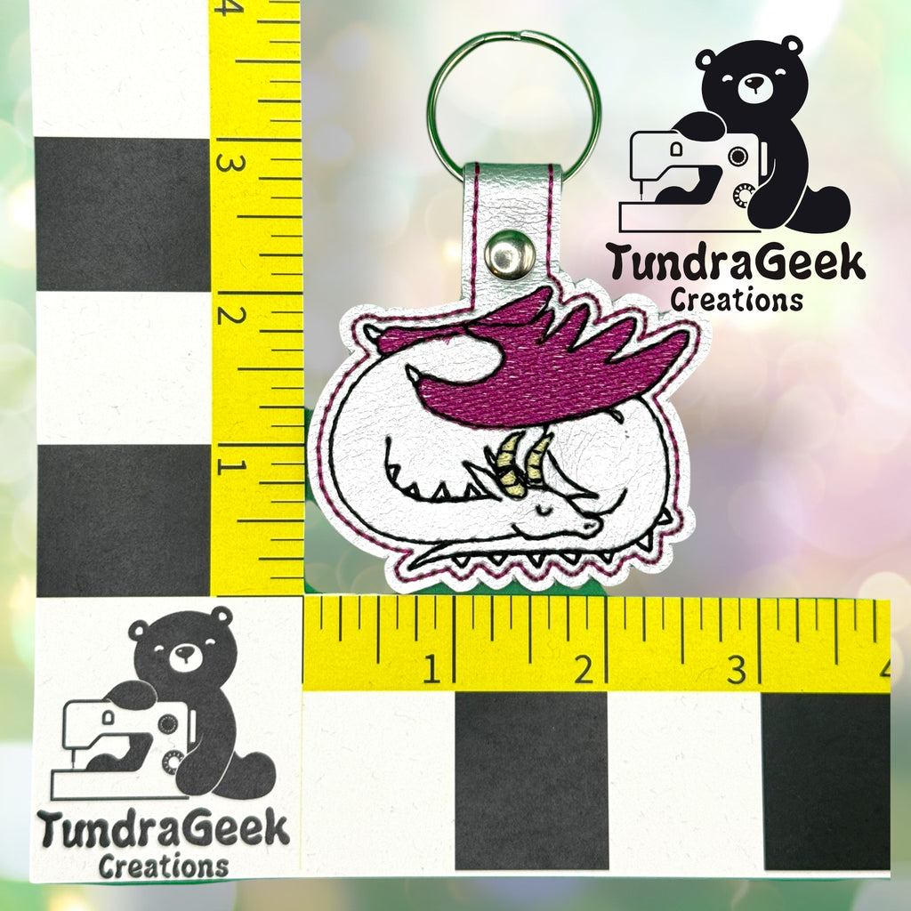 Embroidered keychain featuring a sleeping white dragon with purple wings, displayed next to rulers showing its size, branded with TundraGeek Creations logo.