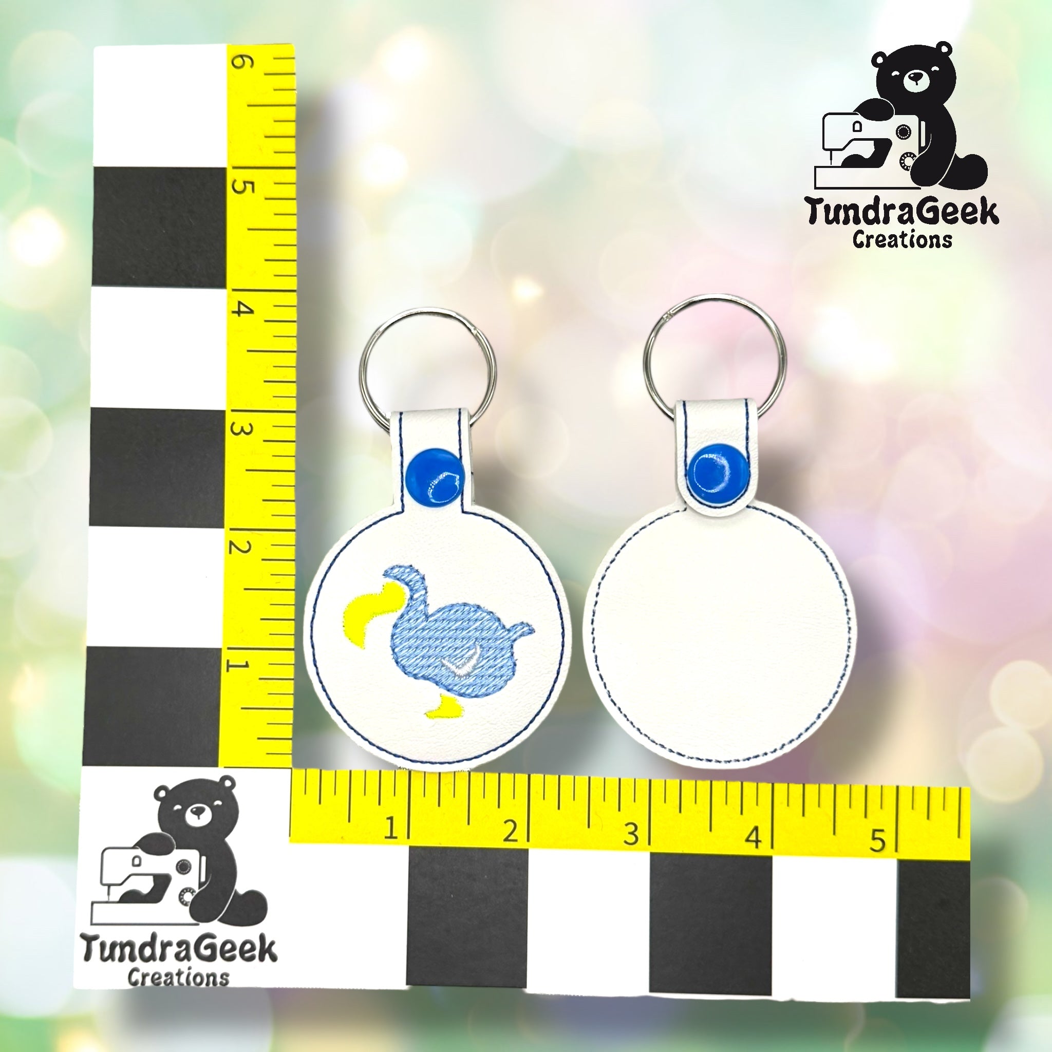 Two round keychains shown front and back; one has a blue dodo bird design. A black and yellow ruler and TundraGeek Creations logo are also visible.