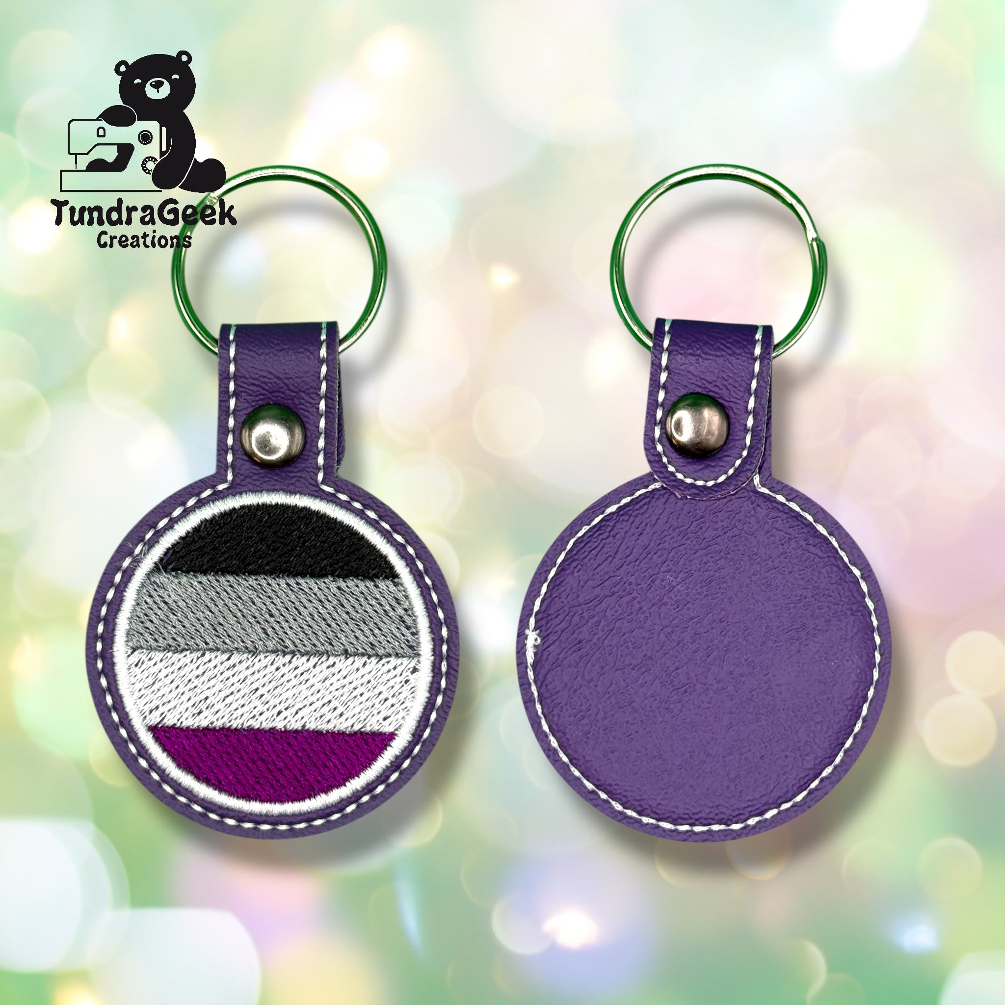 Two purple circular keychains with silver snap buttons and green rings; one displays the asexual pride flag, the other shows the plain back. TundraGeek Creations logo is in the corner.