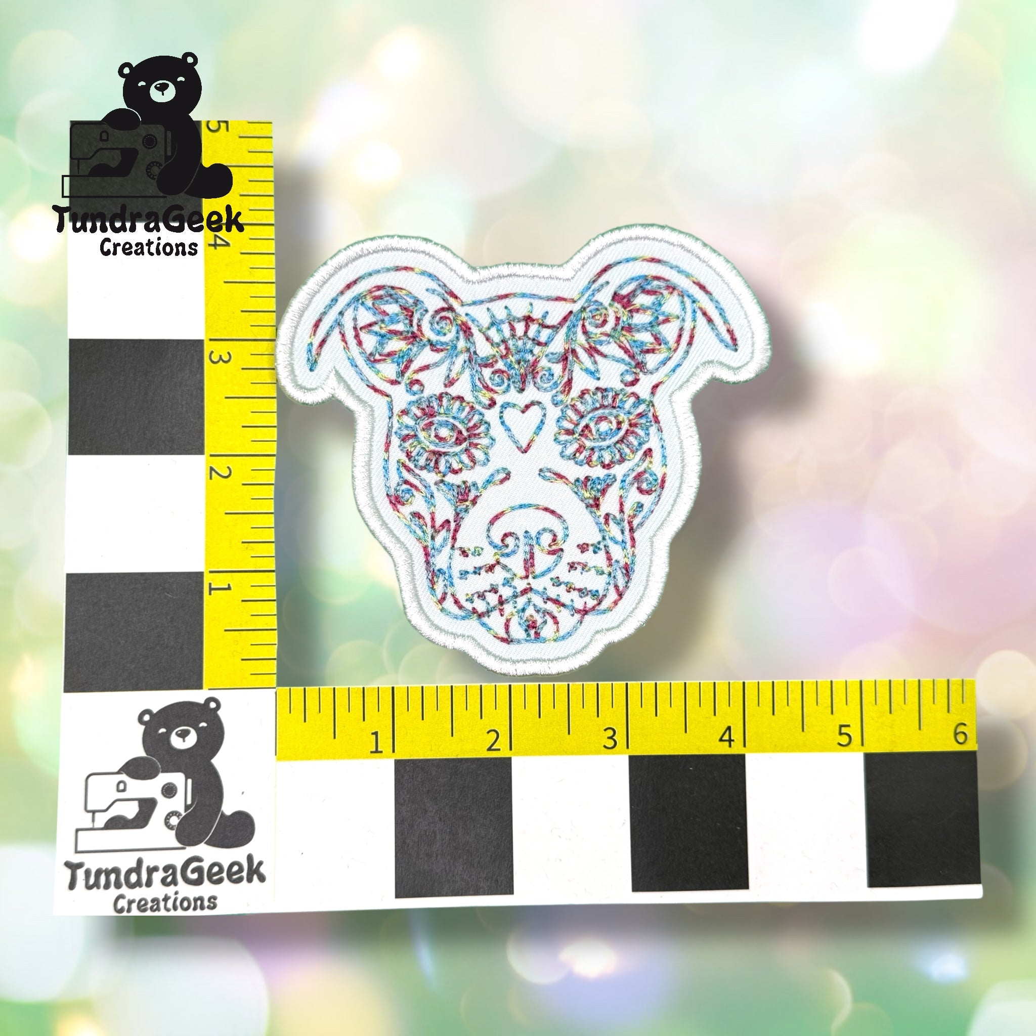 Embroidered patch of a dog’s face with colorful thread, measured against a black, white, and yellow grid ruler marked with TundraGeek Creations logo.