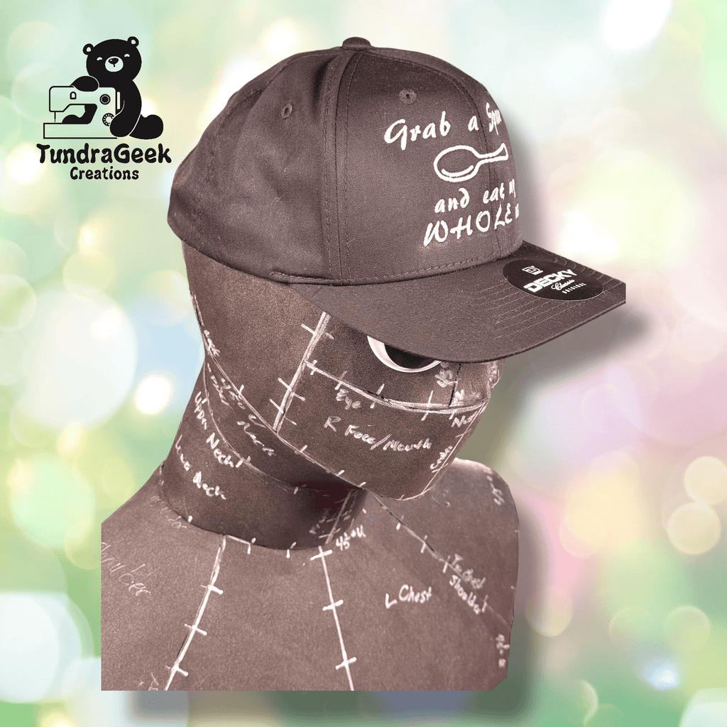 A matte black mannequin head wearing a black baseball cap with white text, against a pastel bokeh background. The TundraGeek Creations logo is in the top left corner.