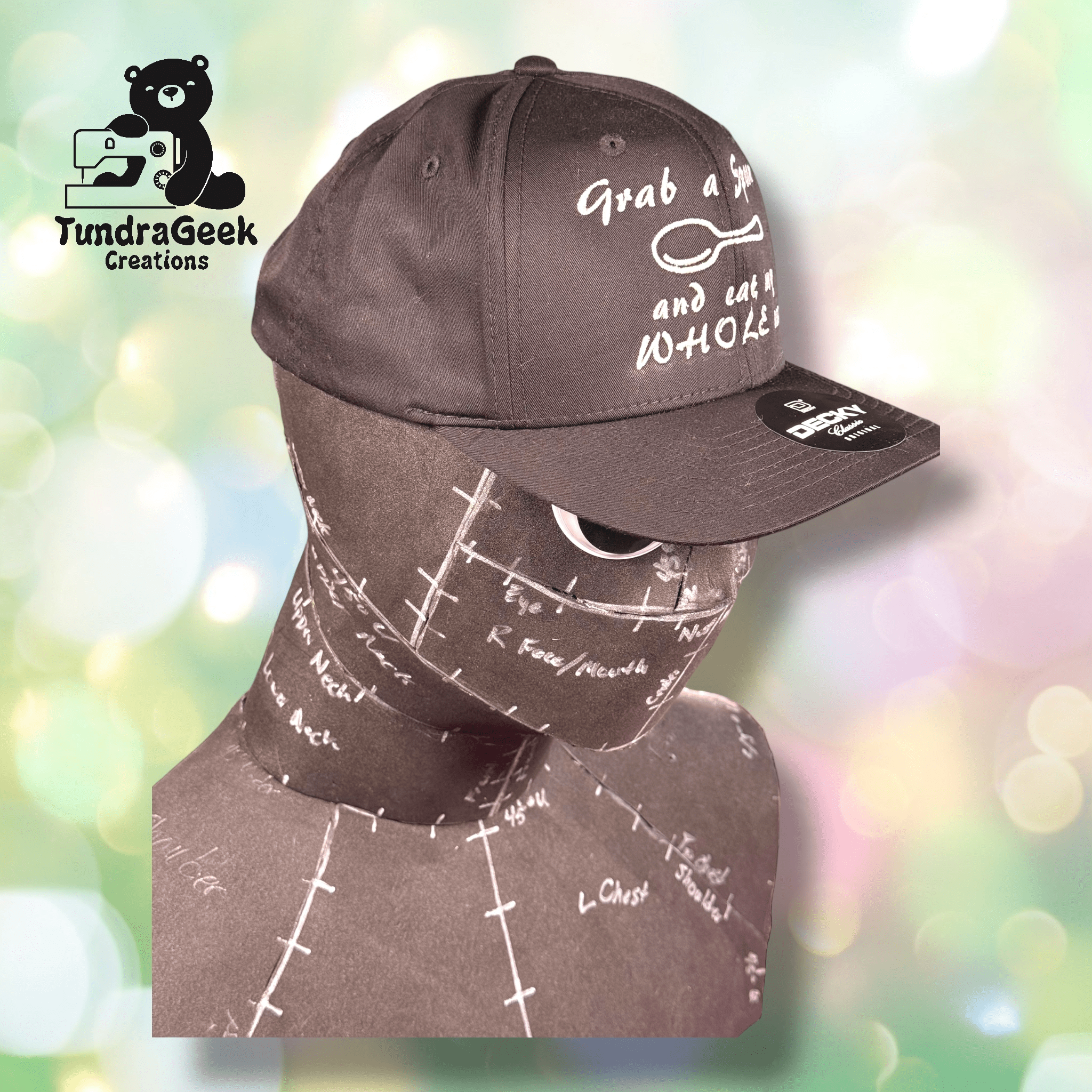 A matte black mannequin head wearing a black baseball cap with white text, against a pastel bokeh background. The TundraGeek Creations logo is in the top left corner.